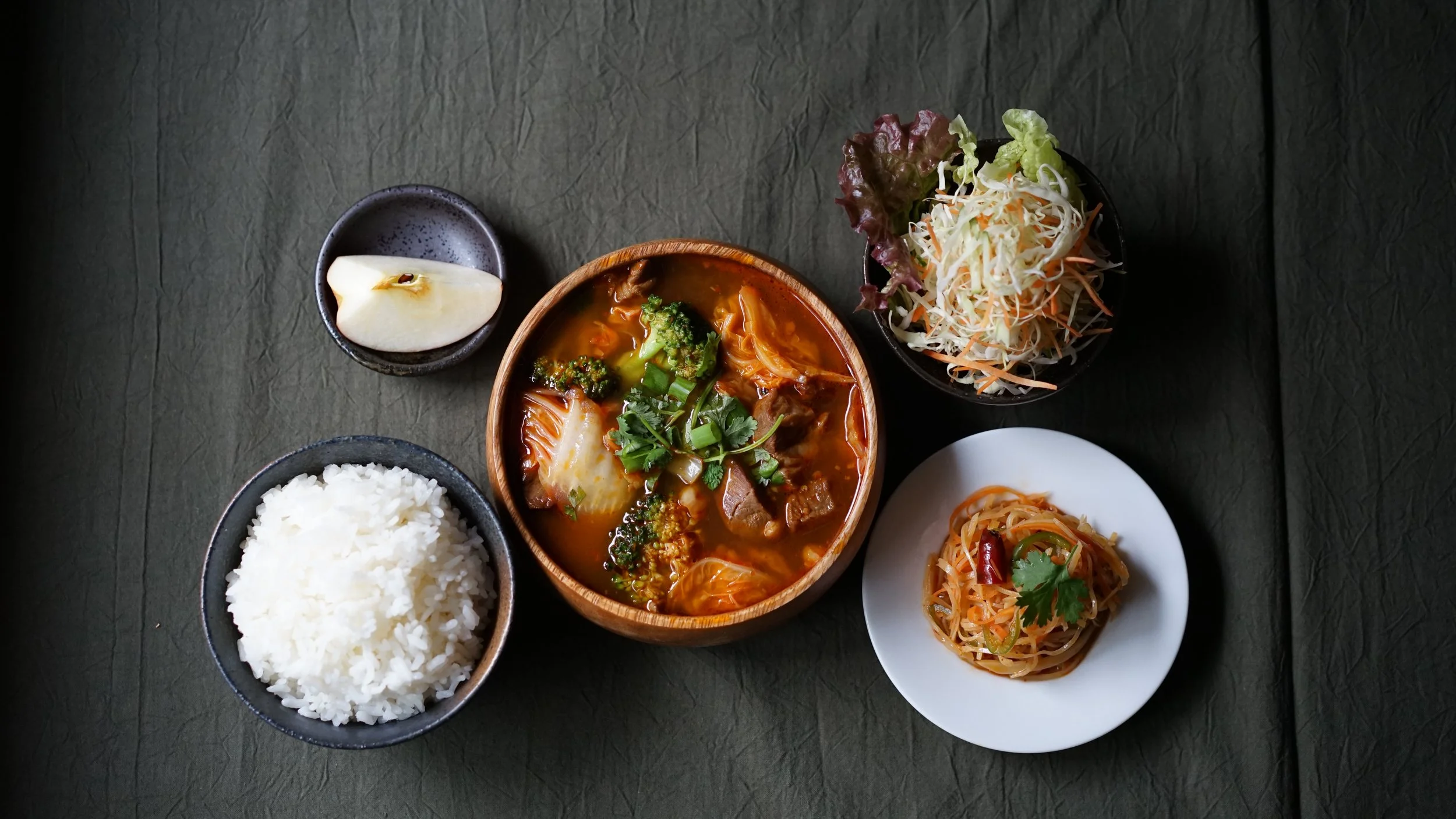 A traditional Asian meal with a bowl of spicy soup with vegetables and beef, a bowl of white rice, a small black dish with a slice of pear, a bowl of Asian-style cabbage salad, and a small white plate of noodle salad garnished with cilantro, all on a dark textured surface.