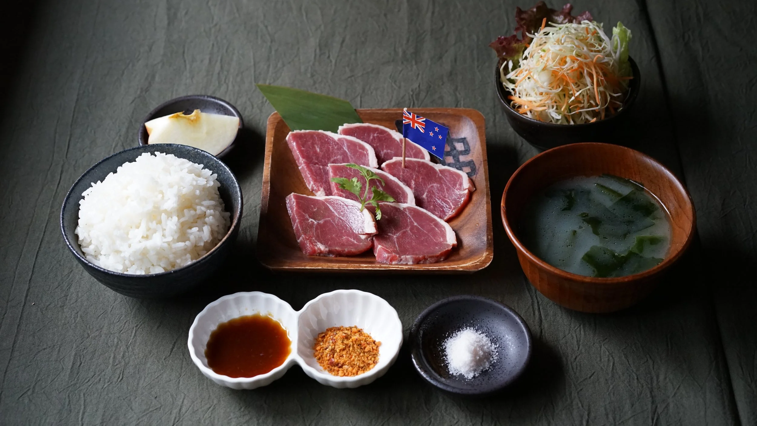 A Japanese meal featuring sliced raw beef, white rice, a small bunch of herbs, dipping sauces, and a bowl of miso soup with seaweed, accompanied by a bowl of shredded vegetables and a slice of apple on a dark green table.