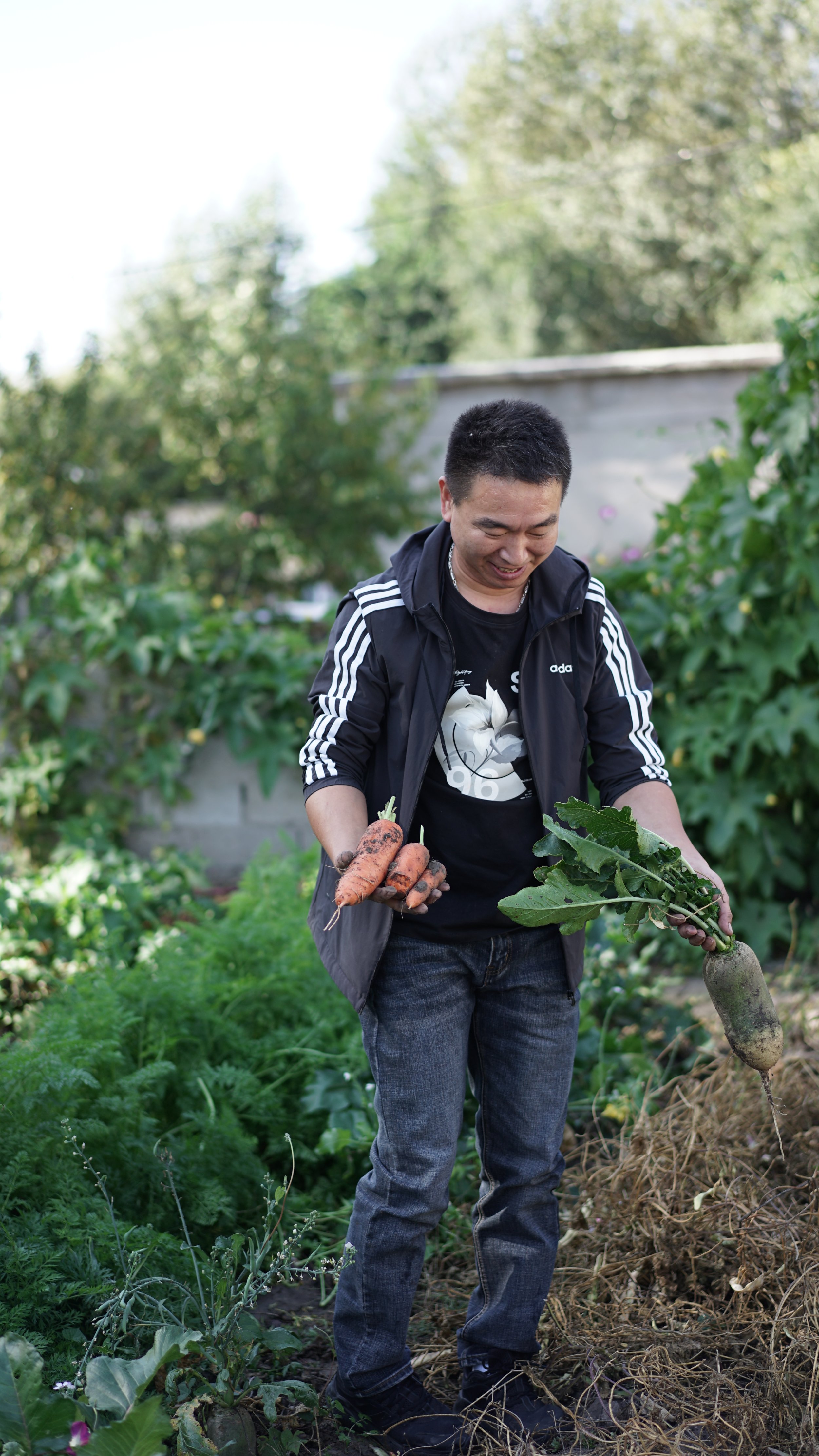 SUYAOLITU is smiling while holding freshly harvested carrots and a large root vegetable in a garden with green plants and trees.
