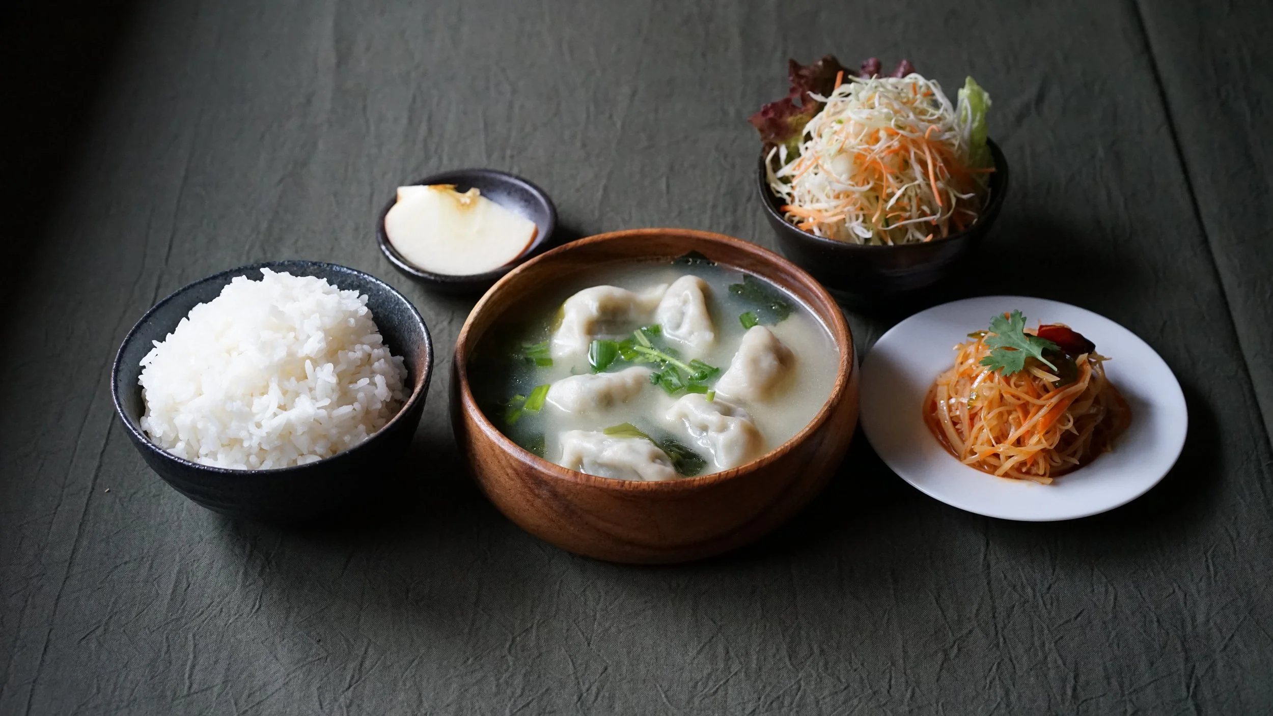 A traditional Asian meal consisting of a bowl of steamed white rice, a bowl of dumpling soup with green onions, a small dish of pickled ginger, and three side dishes: a bowl of shredded cabbage salad and a small plate of shredded carrots garnished with cilantro and chili pepper on a dark table.