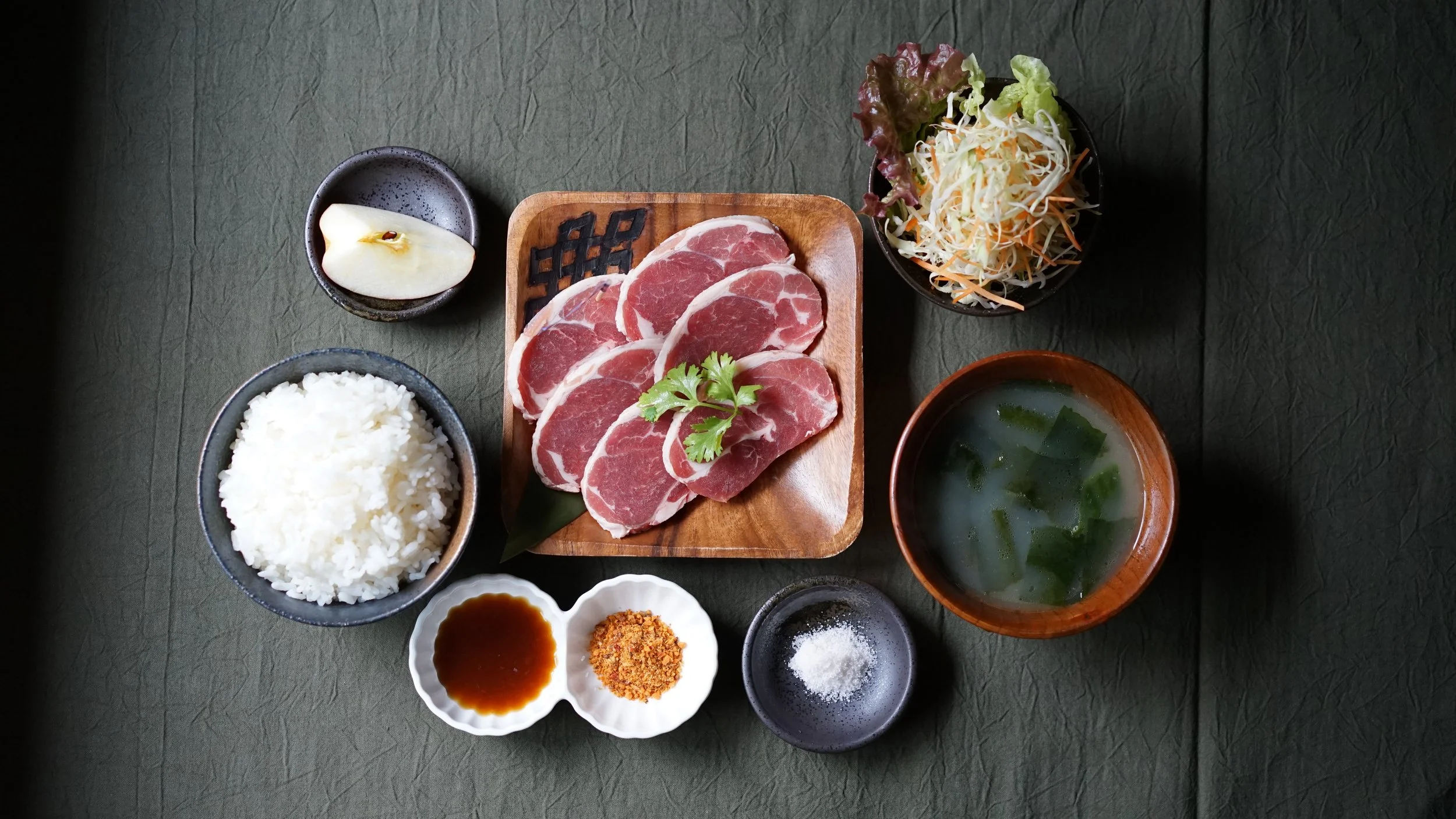 A Japanese-style meal featuring raw sliced meat, steamed rice, salad, miso soup, a pear slice, and condiments including soy sauce and spices on a dark green table.