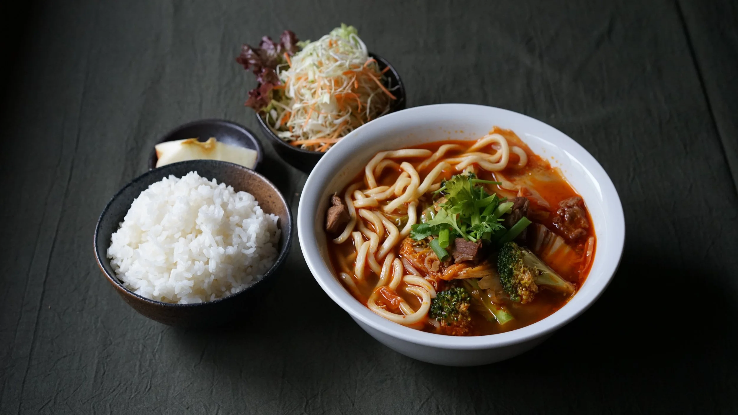 A bowl of ramen noodles with beef, broccoli, and vegetables in a red broth, garnished with cilantro; served with a side of white rice, coleslaw, and pickles on a dark gray table.