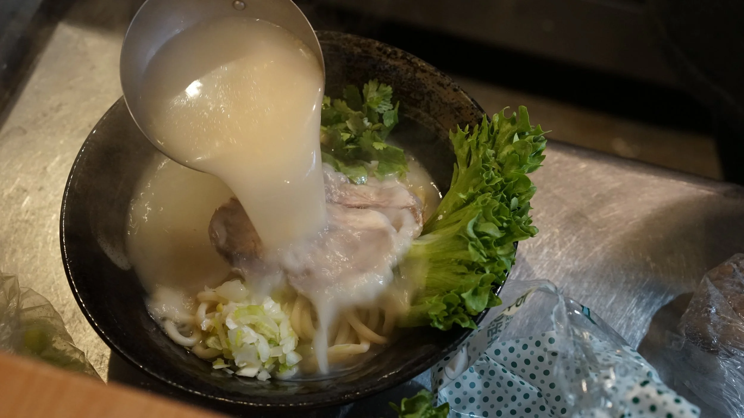 Pouring broth into a bowl of noodles, chicken, lettuce, and chopped green onions.