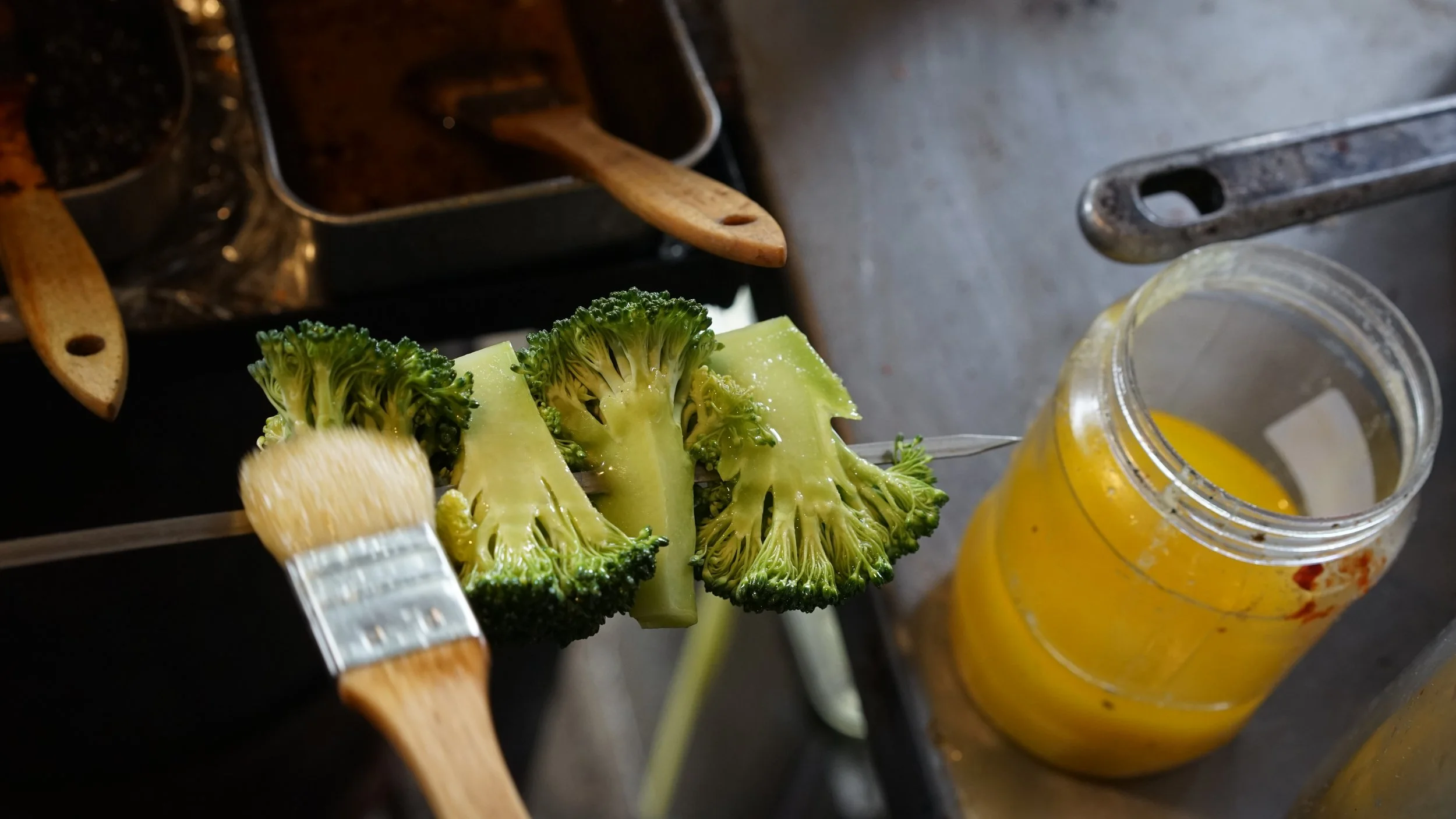 Fresh broccoli being coated with yellow sauce or paint with a pastry brush, near a jar of yellow sauce on a kitchen countertop.