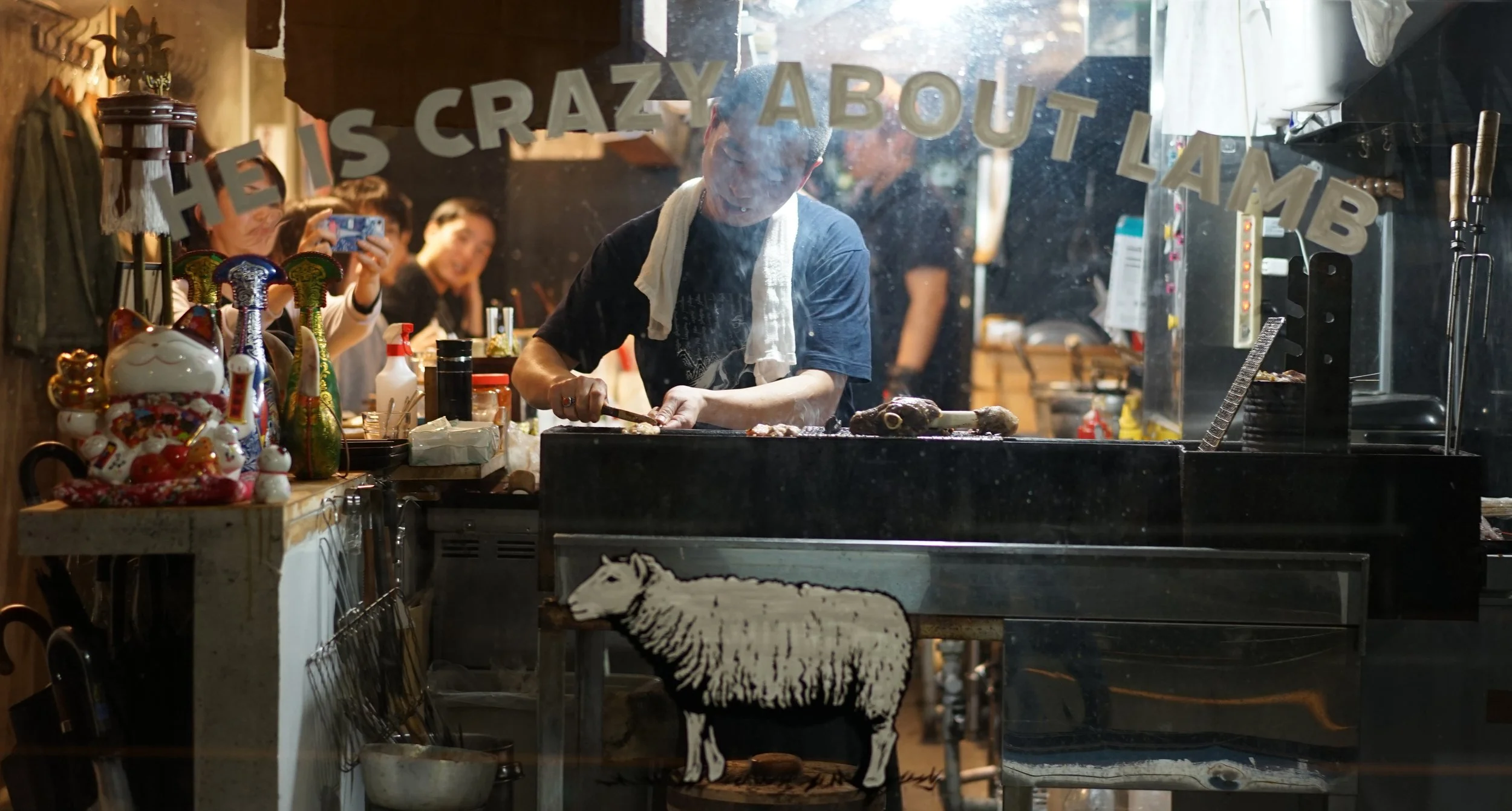 A person cooking meat on a grill inside a restaurant named 'He is Crazy About Lamb,' with customers taking photos and a variety of decorative objects, including a maneki-neko and a sheep illustration, visible inside.