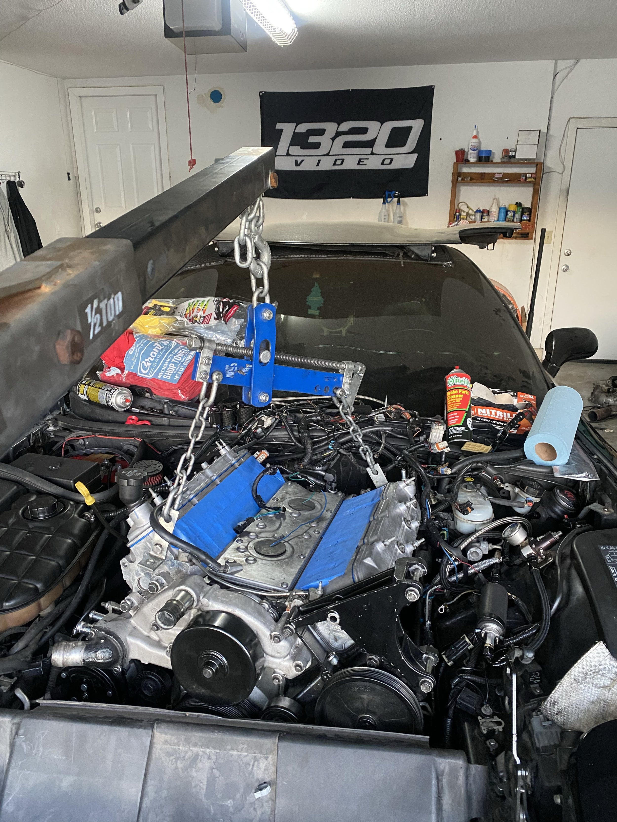 A car in a garage with the hood open and engine being worked on, with engine hoist, tools, and supplies around.