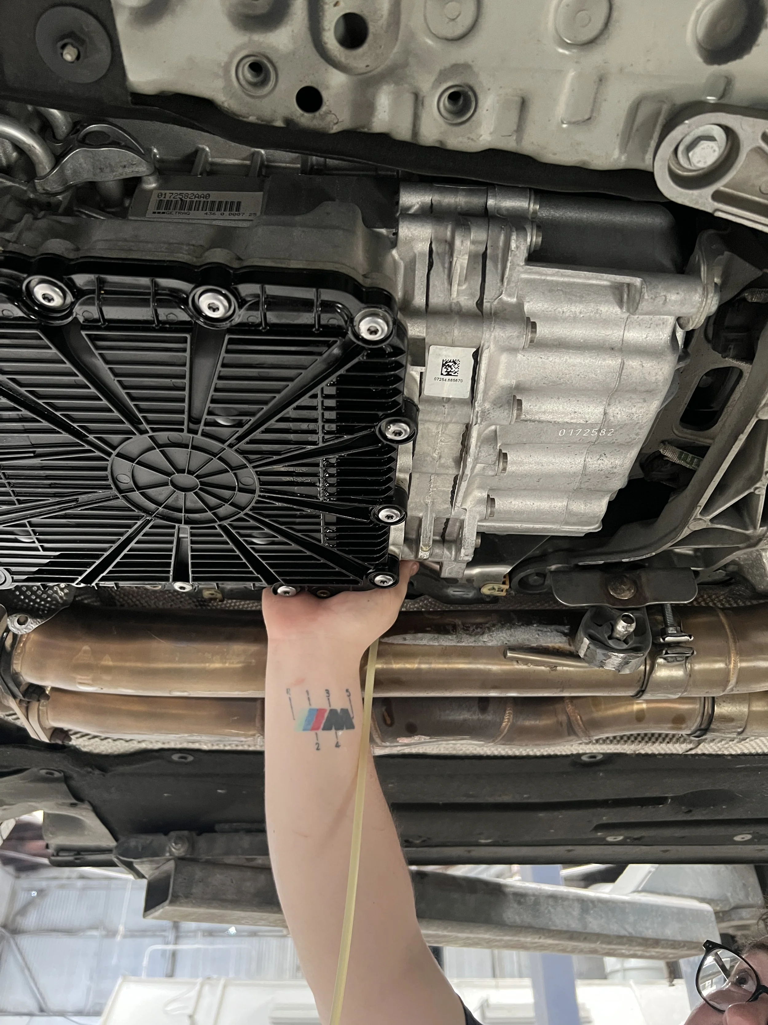A person with a tattoo on their arm inspects or works on the underside of a car engine, holding a component near the engine block and transmission, with exhaust pipes visible below.