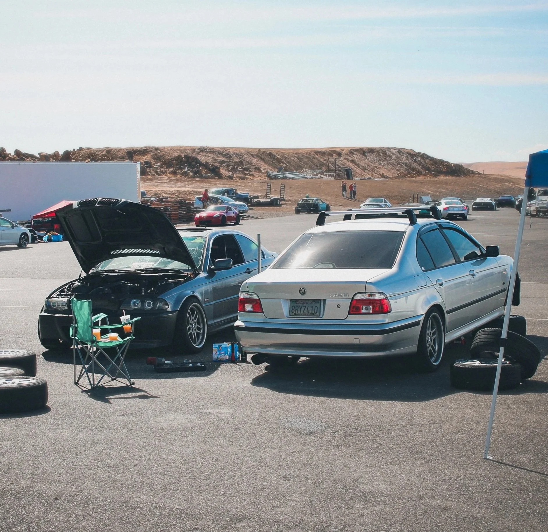 Two cars, one with its hood open, are parked on a race track pit stop area. There is a small table with drinks and snack containers, and a few tires stacked nearby. In the background, there are more cars and a dirt hill.