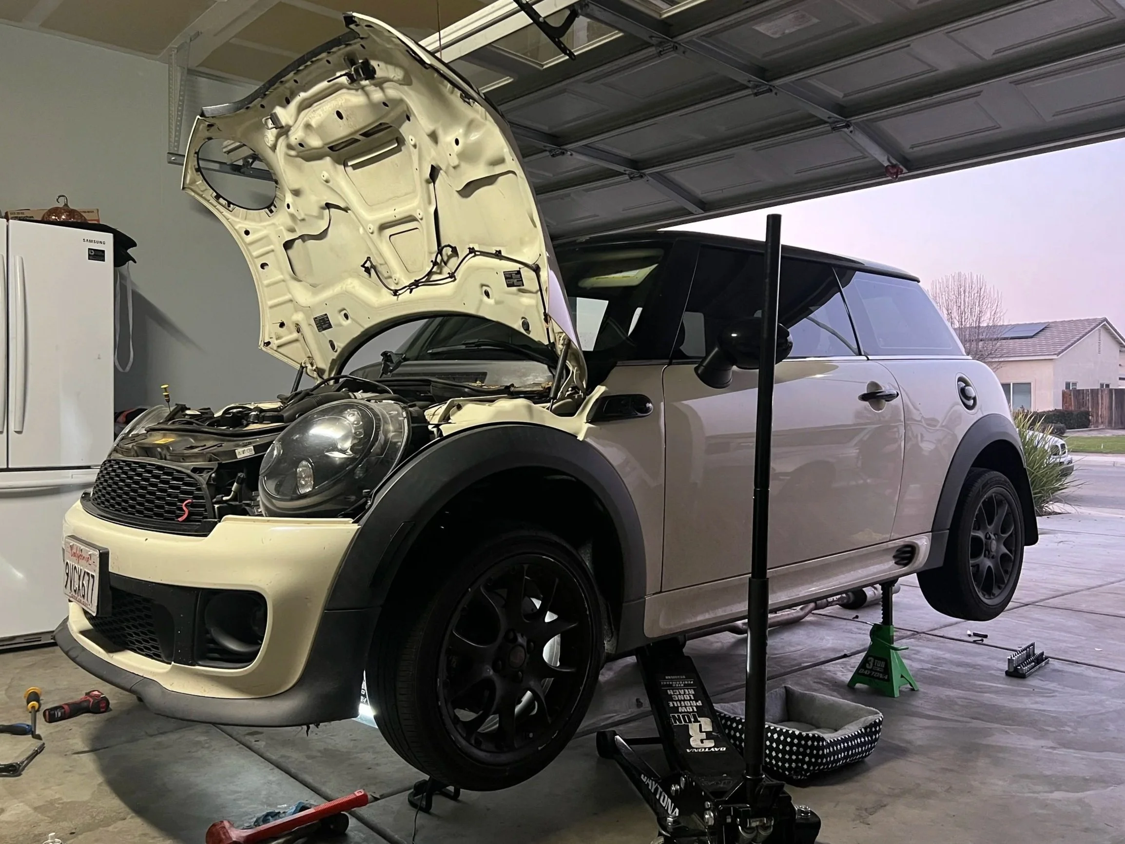 A white Mini Cooper car is lifted in a garage with its hood open, undergoing repair or maintenance. Various tools and equipment are scattered around the garage floor.