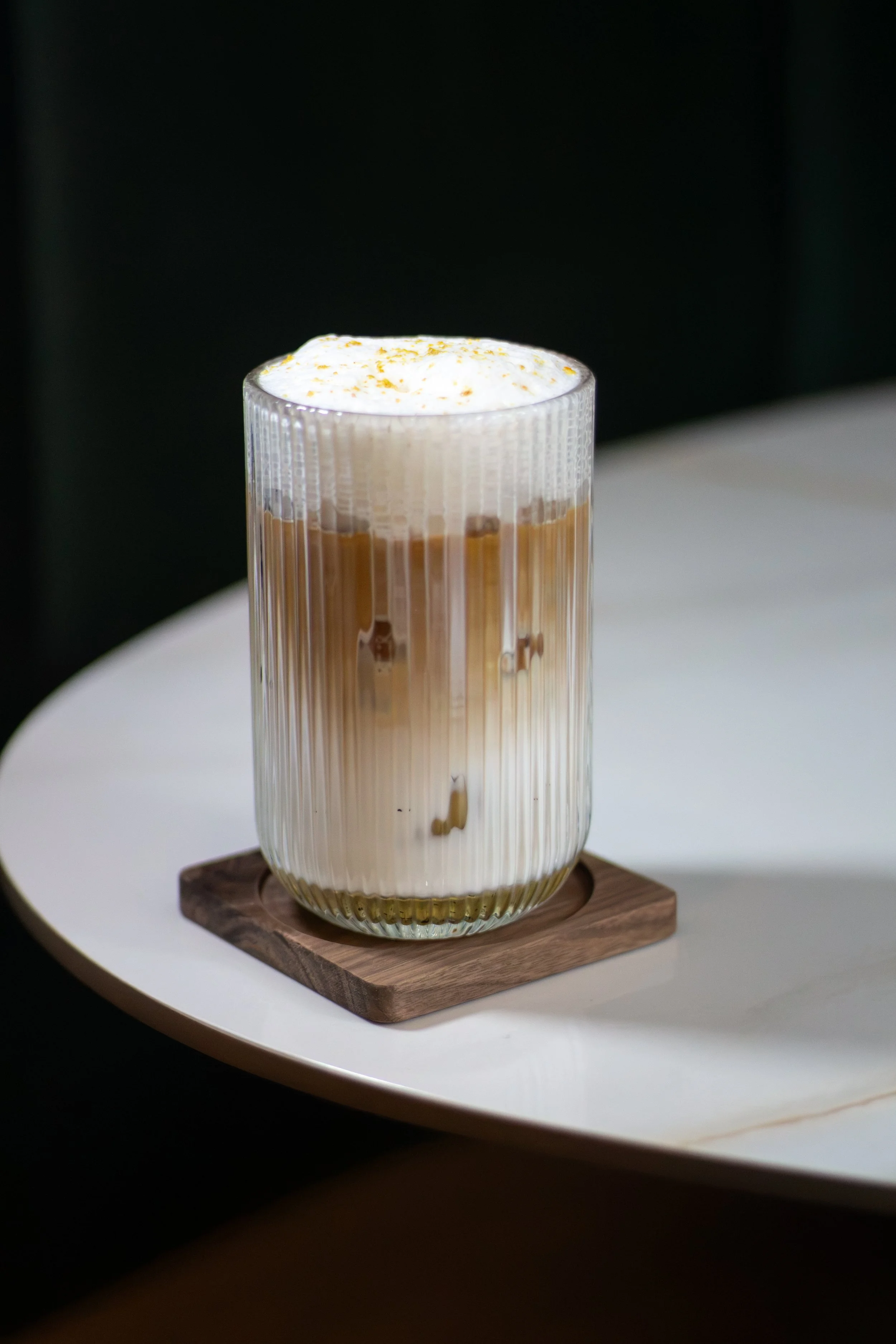 Vaso alto con café con leche y crema batida, sobre una tabla de madera en una mesa blanca.