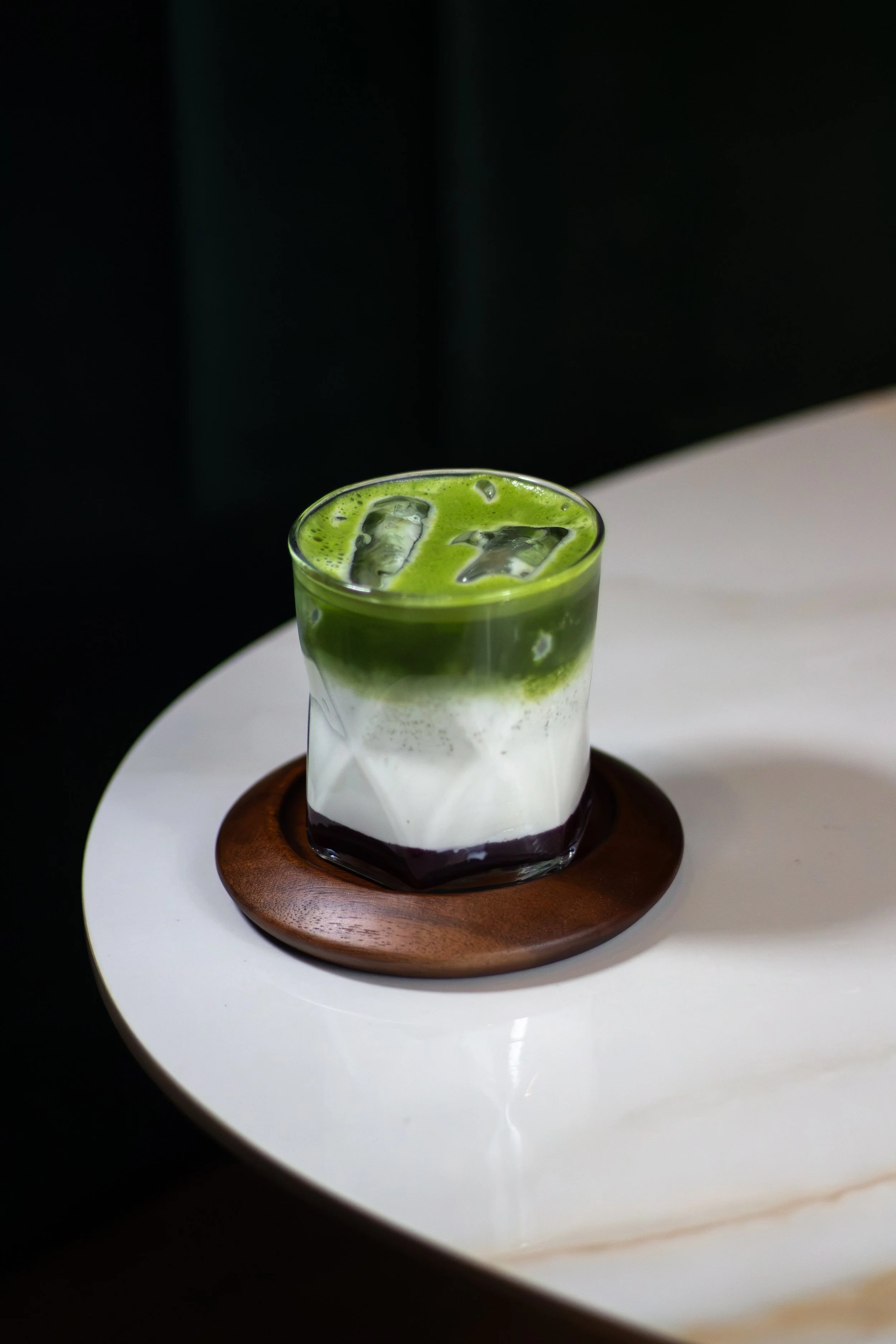 Vaso con bebida de capas verdes y blancas, con hielo, sobre plato de madera en fondo oscuro