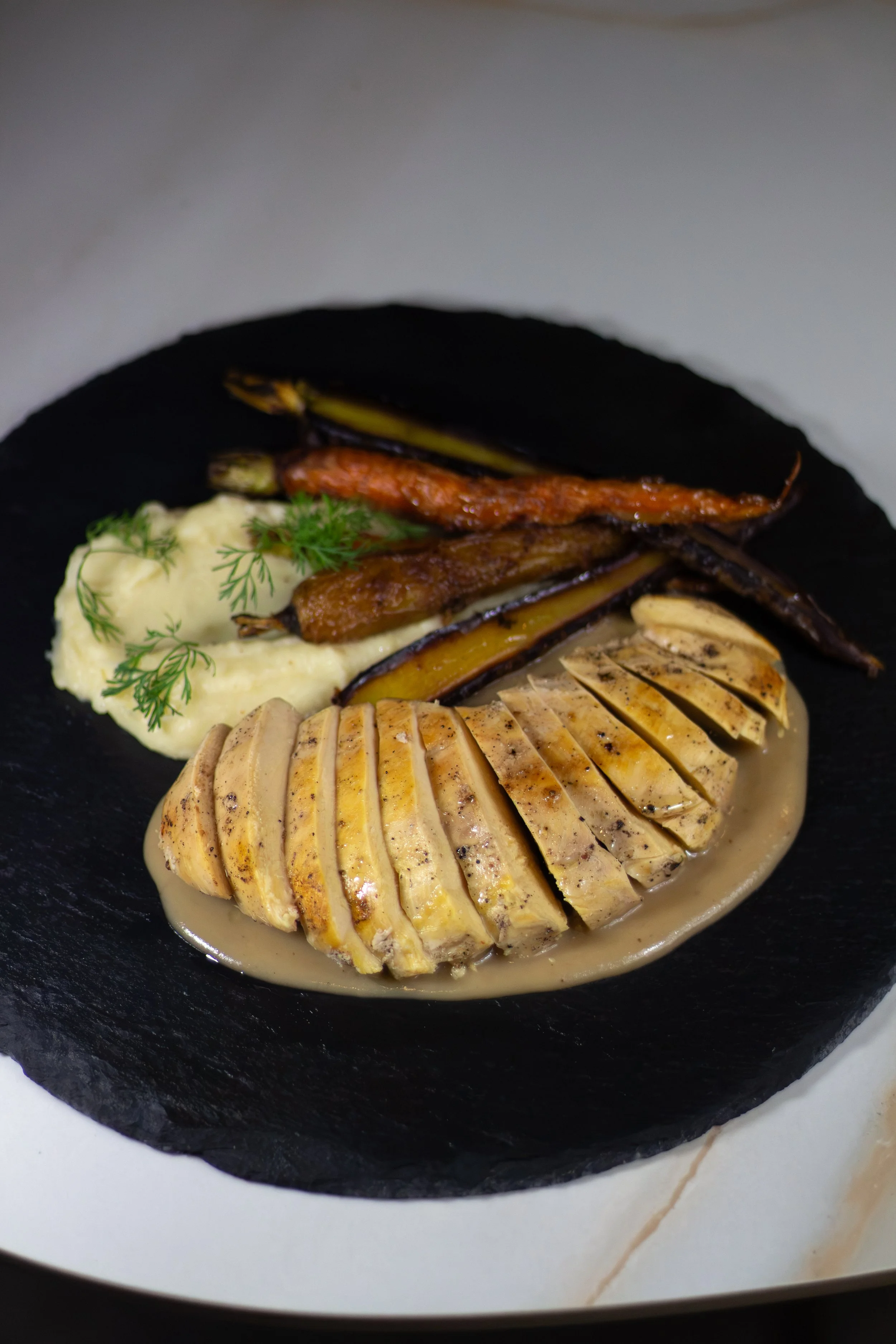 Hombre cortando un pollo relleno y cubierto con salsa, acompañado de puré de papas y verduras asadas, en un plato negro.