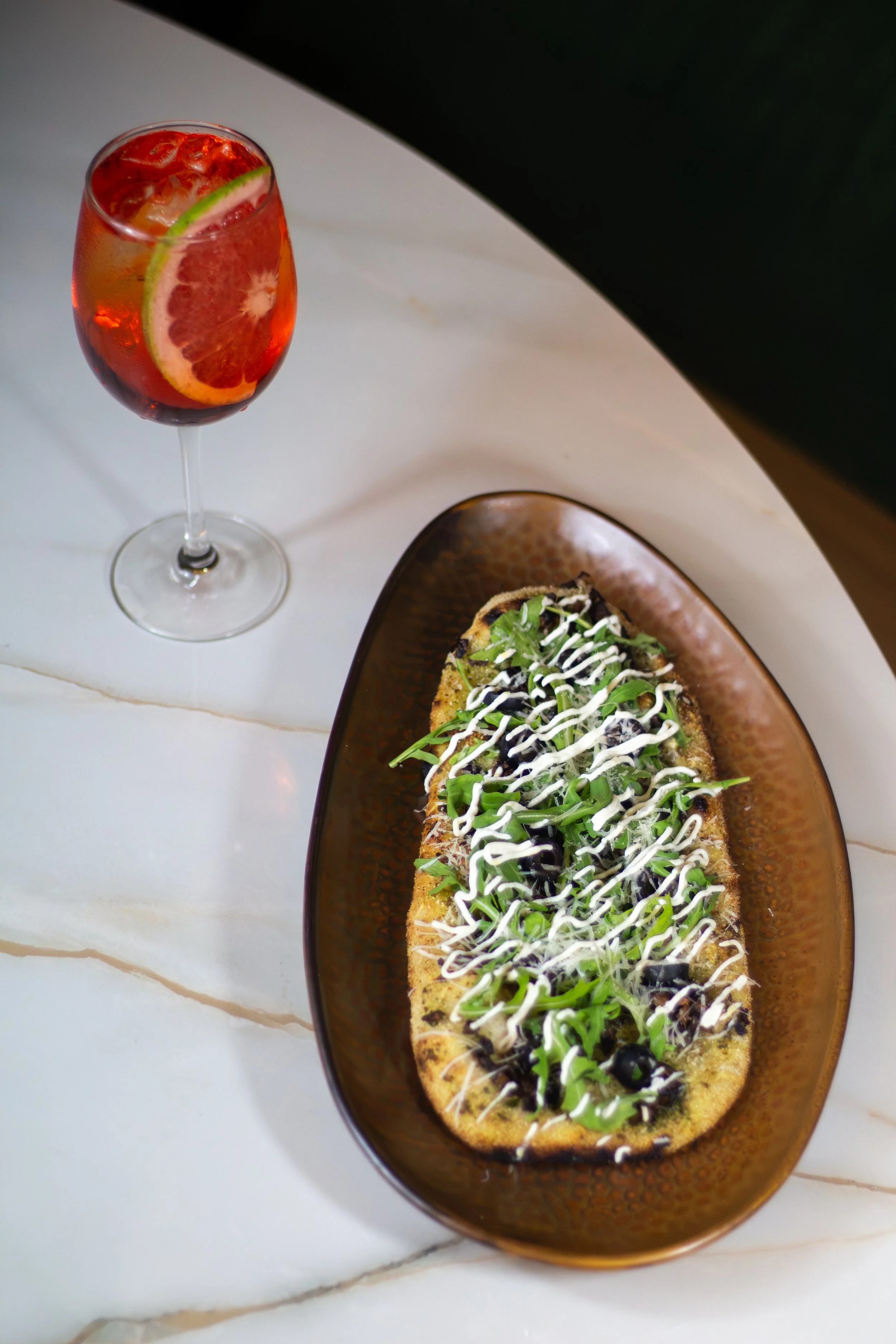 Vaso de cóctel de color rojo con rodaja de lima y hielo y una pizza con queso, aceitunas negras, jalapeños, y verduras en un plato de cerámica.