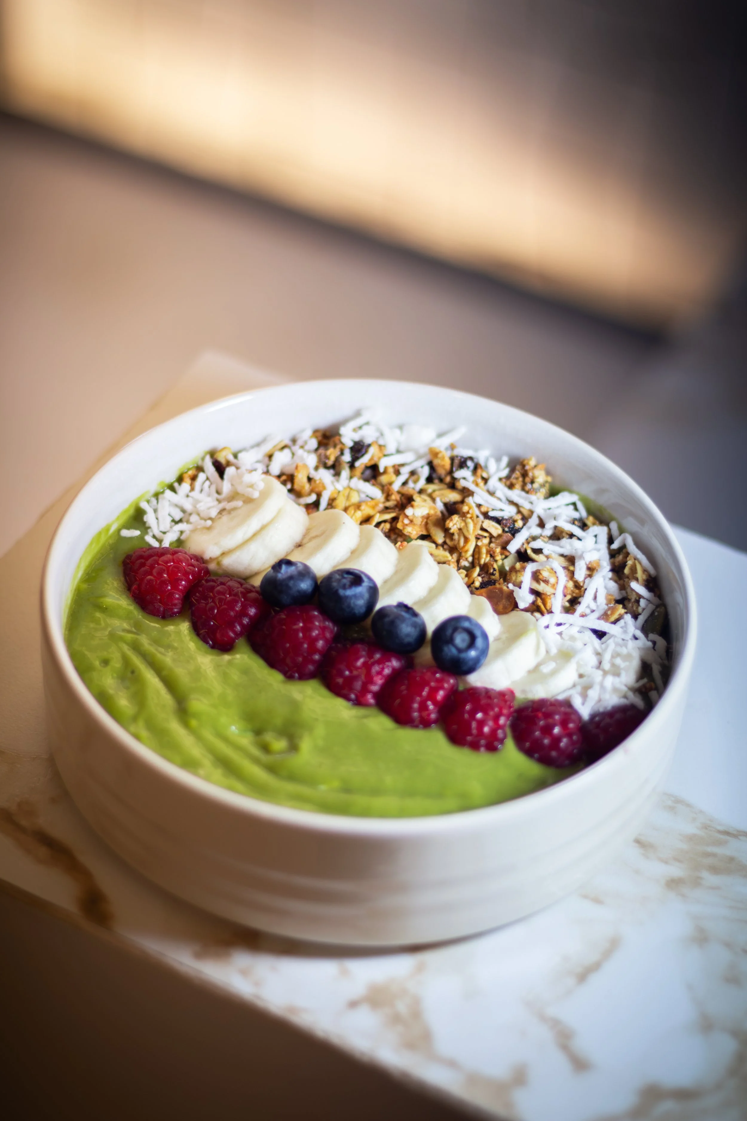 En un recipiente blanco hay un batido o smoothie con frambuesas, arándanos y plátano en la parte superior, decorado con granola y ralladura de coco, sobre una superficie de mármol.