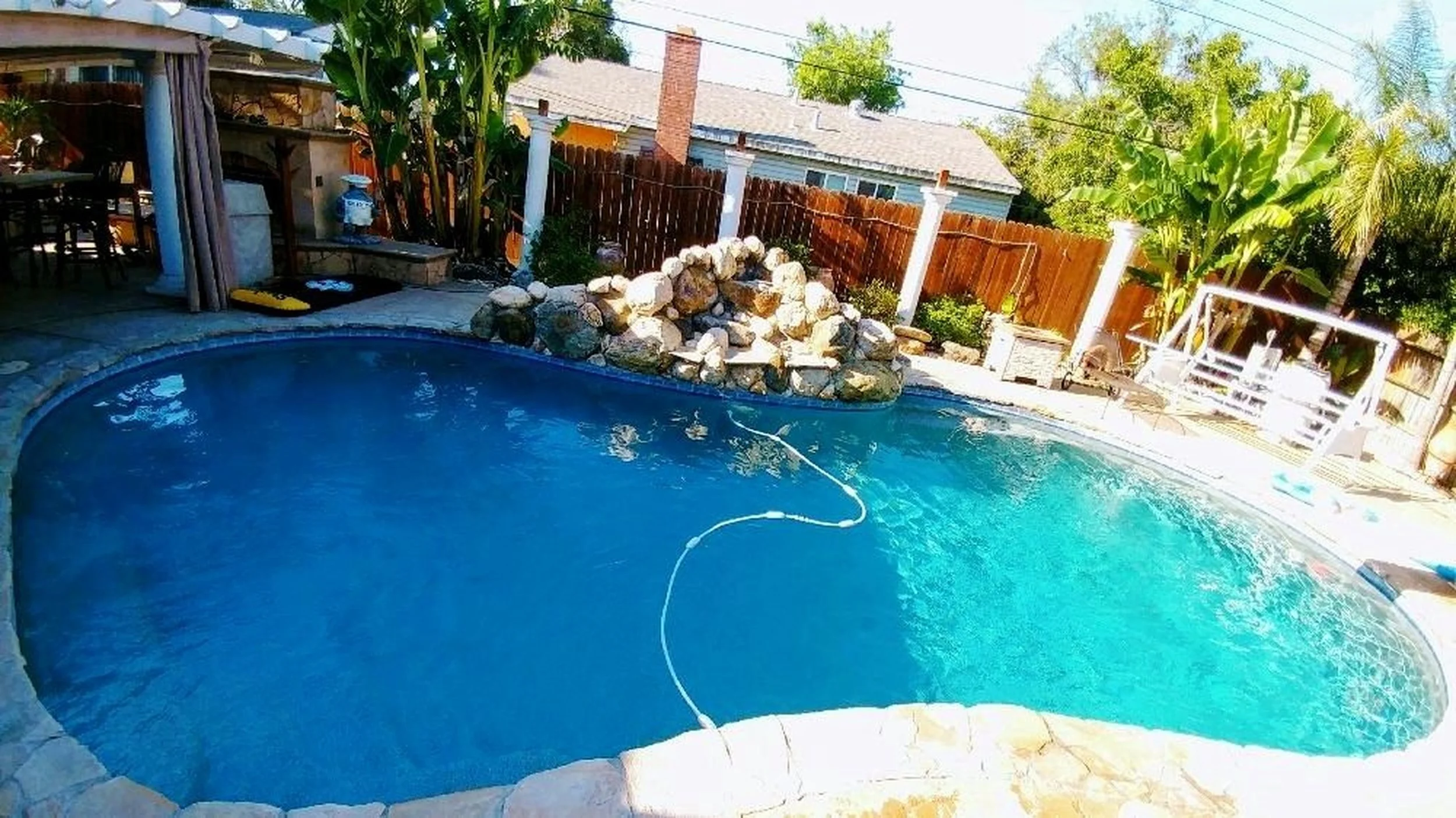 Backyard with a swimming pool featuring a rock waterfall, surrounded by trees and plants, enclosed by a wooden fence, with outdoor furniture and a small covered patio area.