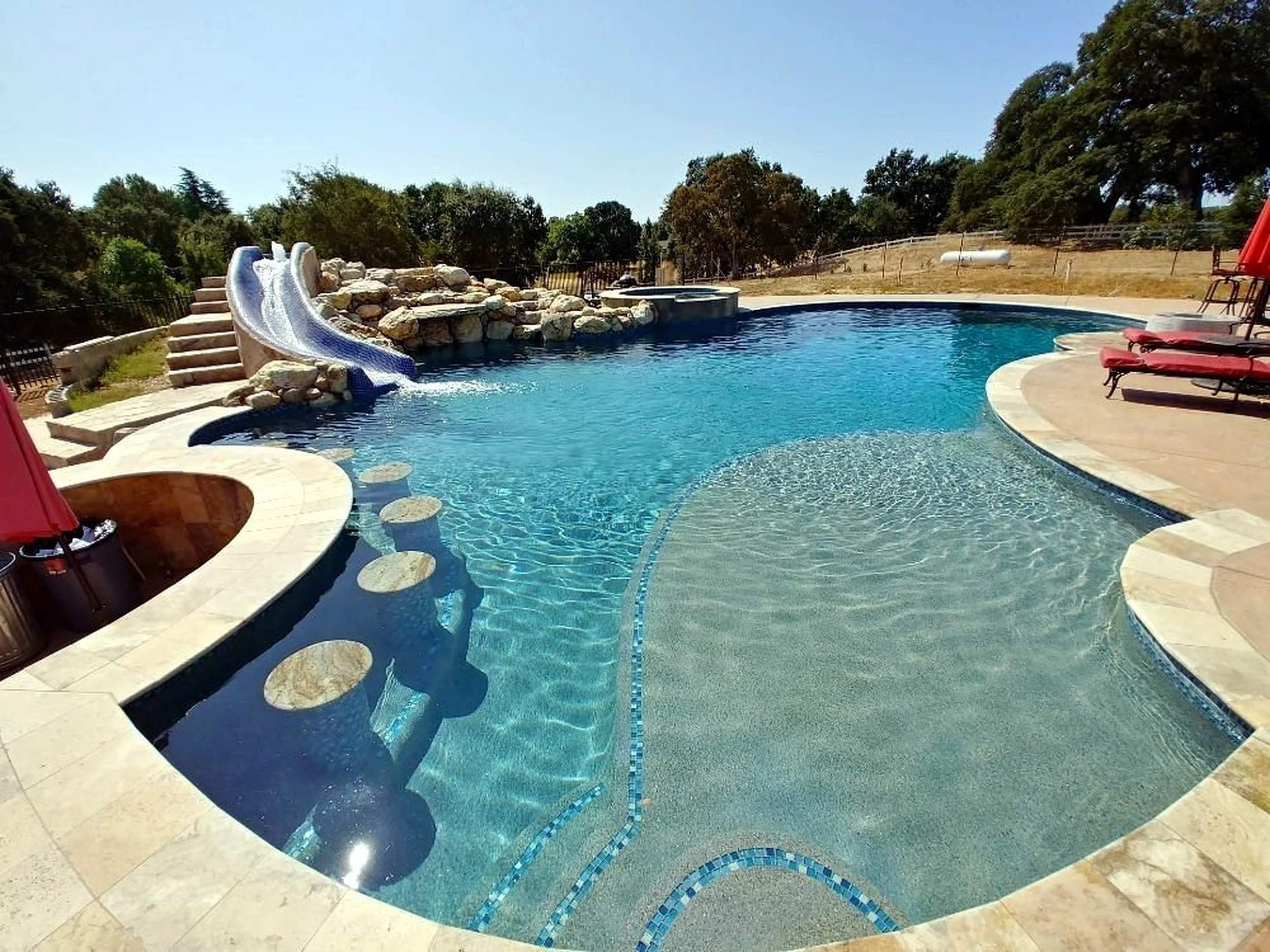 A backyard swimming pool with a small water slide made of rocks, lounge chairs with red cushions, and surrounding trees under a clear blue sky.
