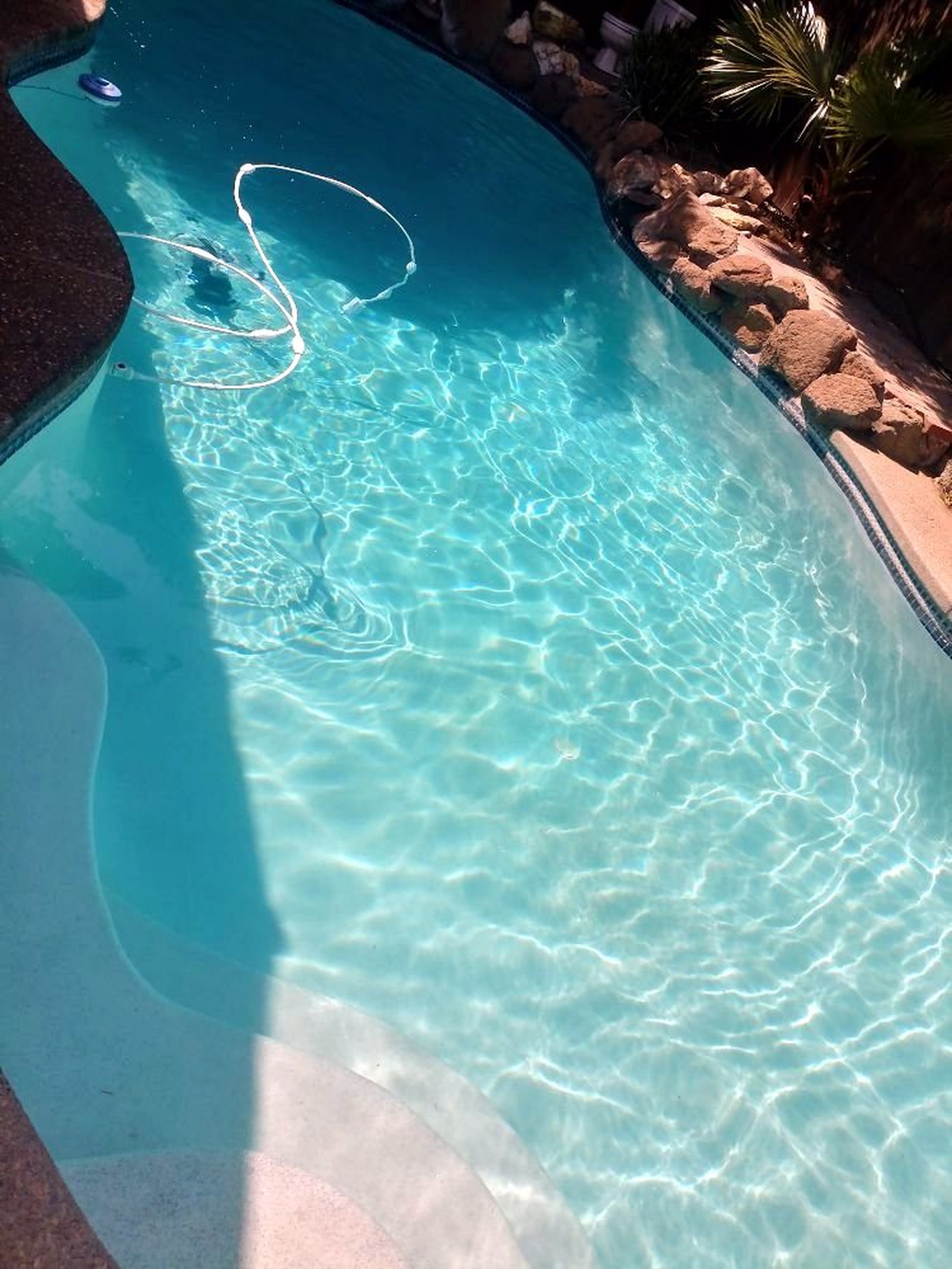 Clear blue swimming pool with sunlight reflections, surrounded by rocks and some trees in the background.