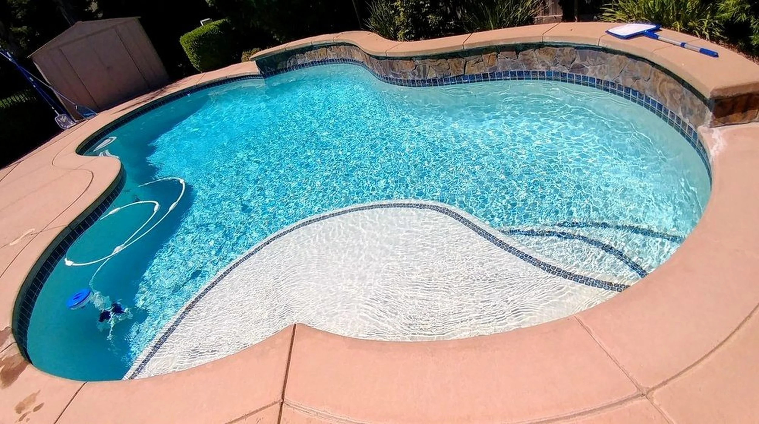 A backyard swimming pool with two sections, one shallower and one deeper, surrounded by a pink concrete deck. There are pool cleaning tools and a fountain on the deck, and a storage shed and lush greenery in the background.