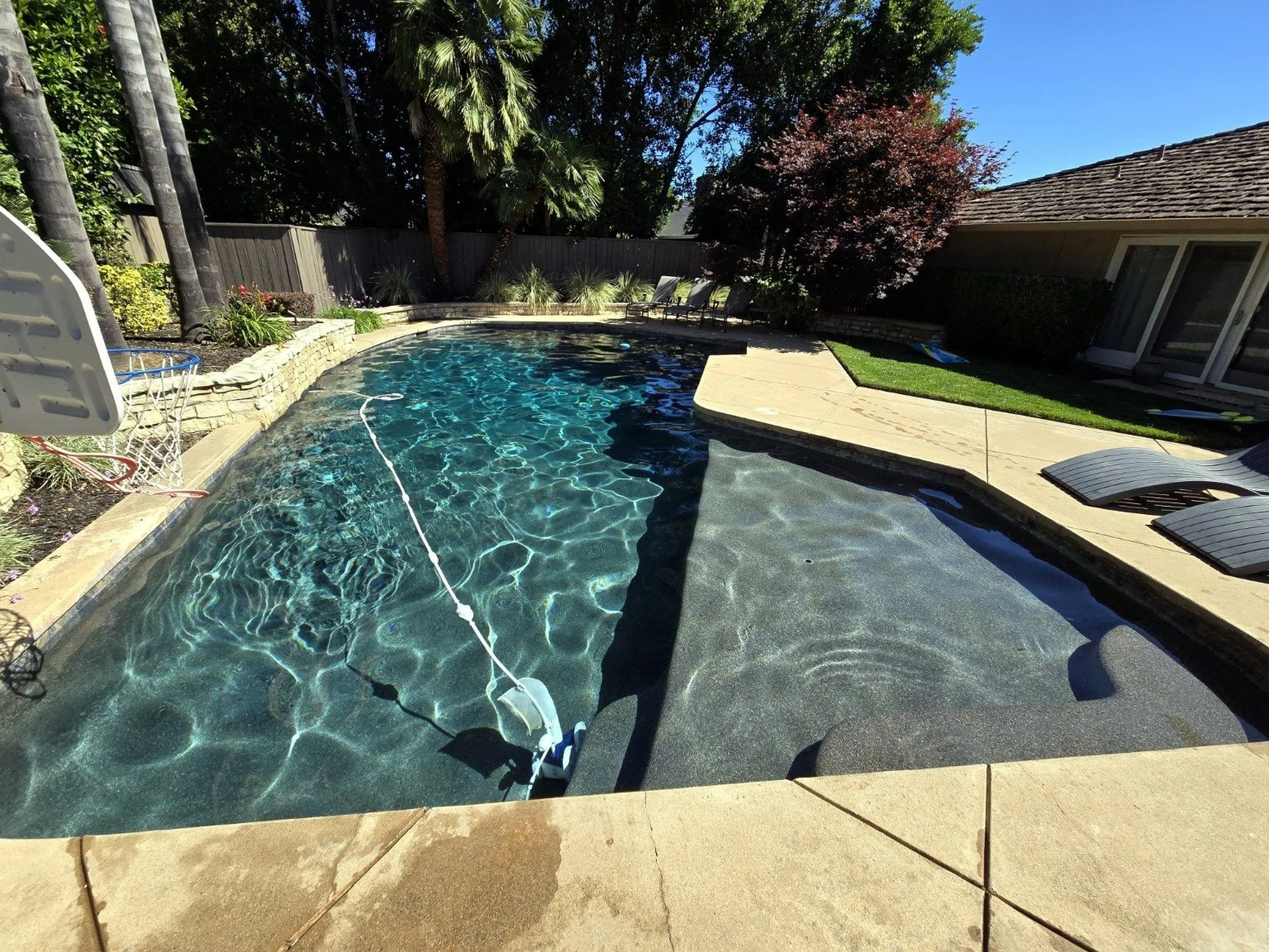 A backyard swimming pool with a pool cleaning device floating in the water, surrounded by a stone deck, lounge chairs, lush trees, and a house with sliding glass doors.