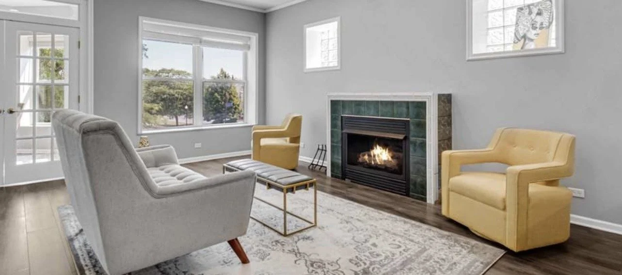 Living room with a fireplace, a gray couch, two yellow armchairs, a small gold coffee table, and large windows