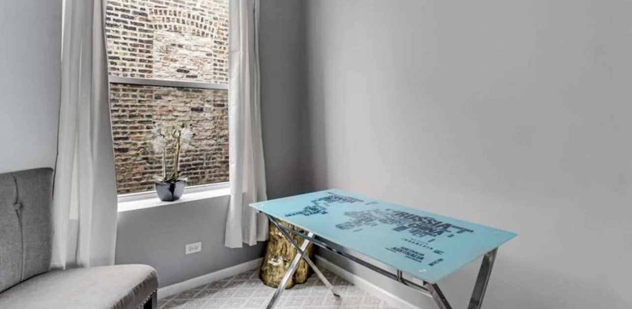 Room with a window, white curtain, potted plant on windowsill, gray sofa, and a folding table with blue surface and black writing, leaning against the wall.