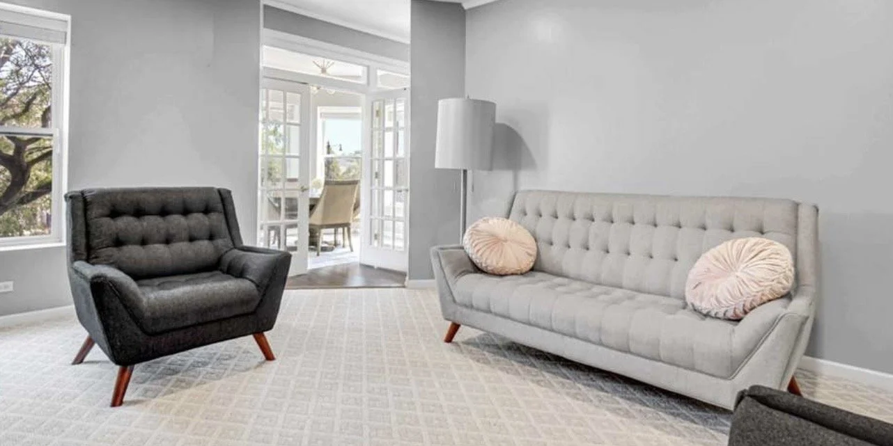 Living room with gray upholstered sofa and dark gray armchair, beige and pink round pillows, gray walls, carpeted floor, and a sunlit dining area visible through glass doors in the background.