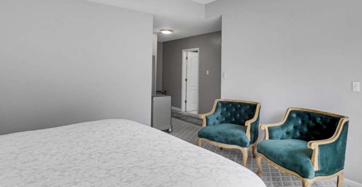 A bedroom with a bed, two teal velvet armchairs, gray walls, and a doorway leading to a bathroom.