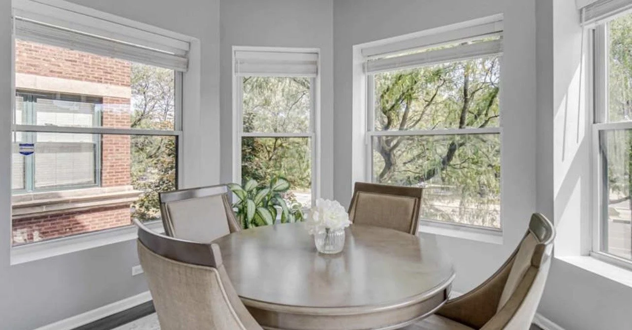 Sunlit dining room with a round table and four upholstered chairs, a centerpiece of white flowers, and multiple large windows revealing trees and a brick building outside.