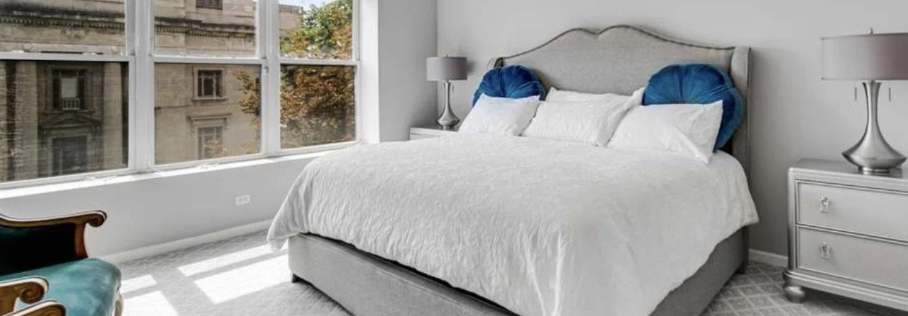 A bedroom with a large bed, white bedding, two blue cushions, and two bedside lamps, illuminated by large windows showing city buildings outside.