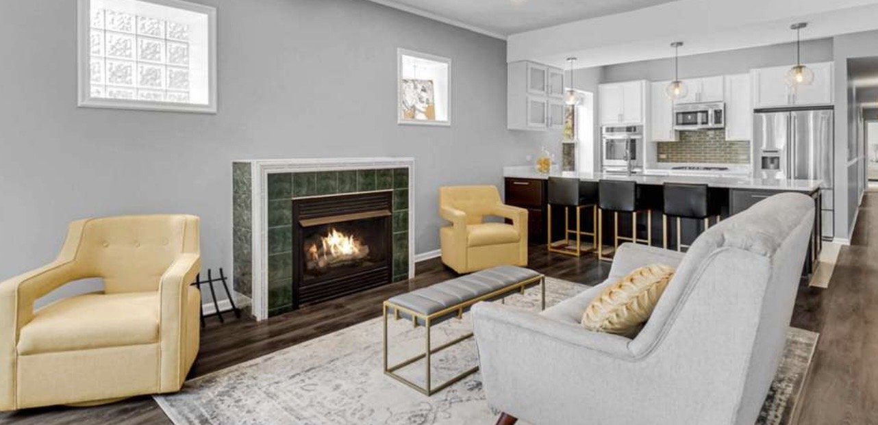 Living room with a lit fireplace, yellow and gray armchairs, a gray sofa with a yellow pillow, a glass-top bench, and views into a modern kitchen with white cabinetry and stainless steel appliances.