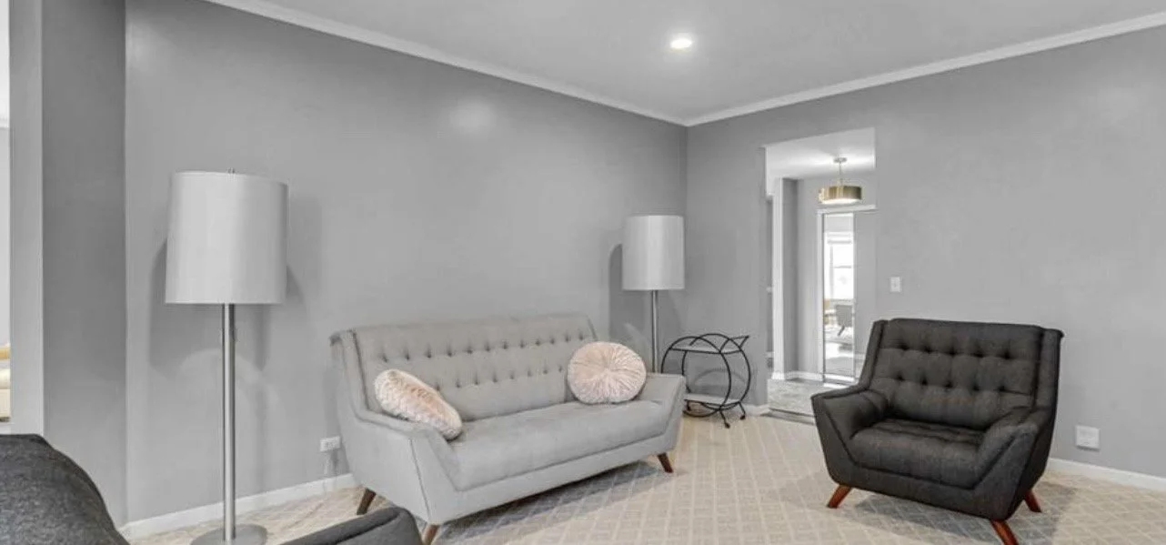 Living room with gray walls, two floor lamps, a light gray sofa with two round pillows, a dark gray armchair, a round side table, and a doorway leading to an outdoor space.