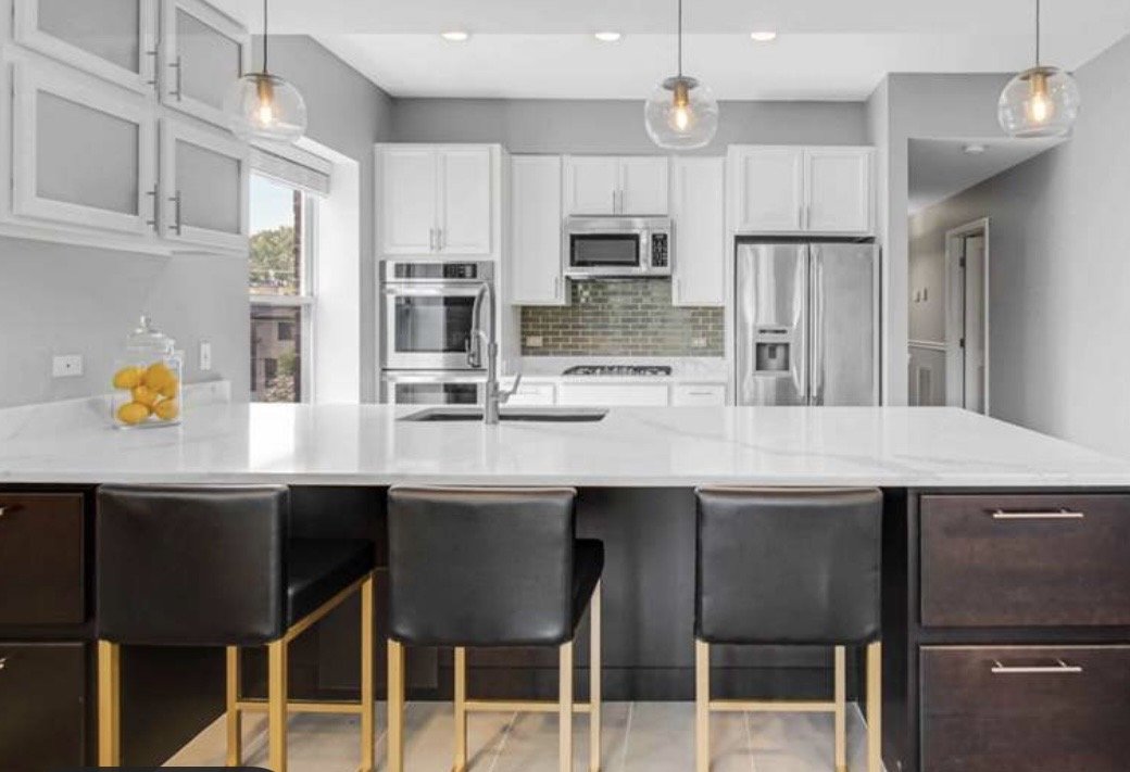Modern kitchen with white cabinets, stainless steel appliances, a large white countertop island with black chairs, pendant lighting, and a gray wall with a small window.