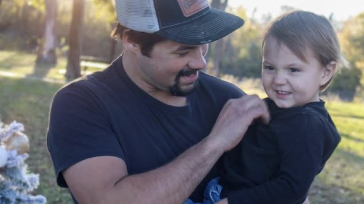 A man and a young girl outdoors, smiling and playing together, with trees and a grassy area in the background.