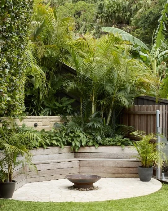 Backyard garden patio with lush tropical plants, potted palm trees, wooden retaining wall, fire pit, and a metal fence