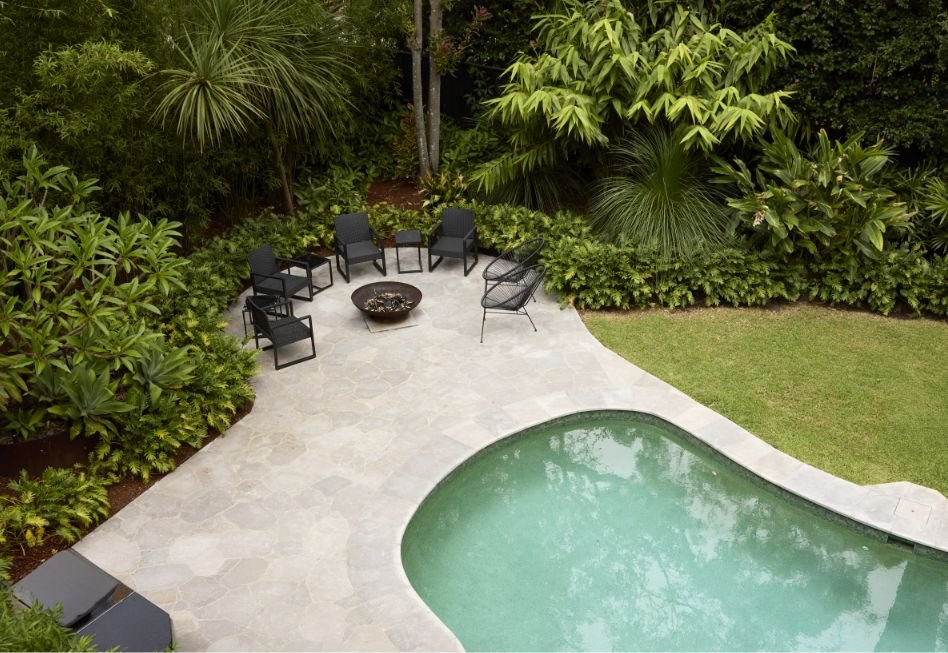 A backyard with a small swimming pool, surrounded by lush green plants and trees, and a patio area with four black chairs and a fire pit.