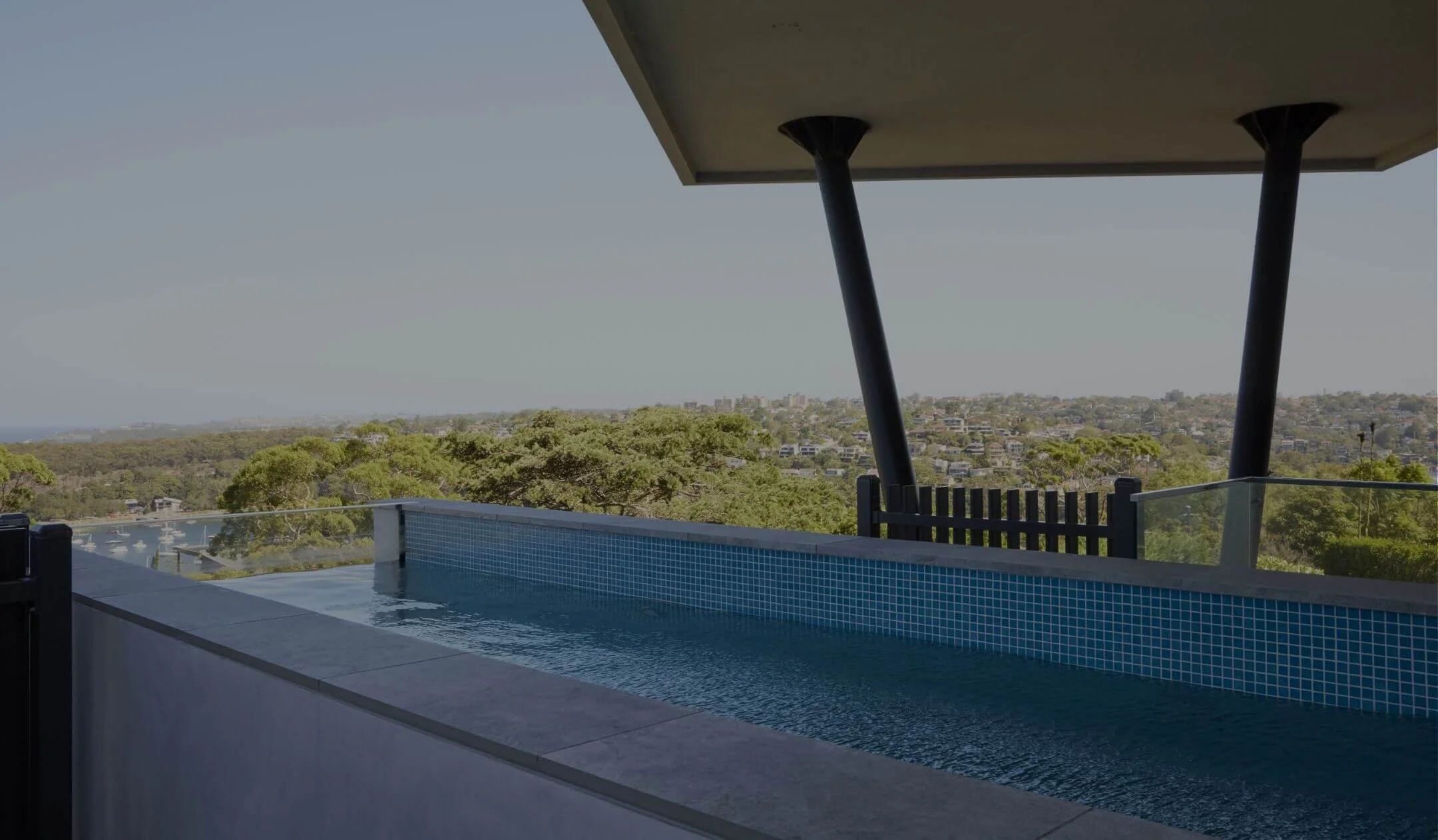 Balcony with a small infinity pool overlooking a cityscape with trees and water in the distance, under a shaded area supported by black angled beams.
