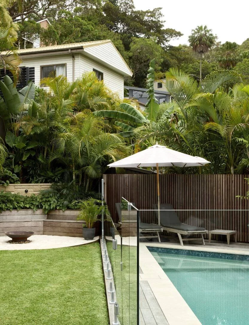 Backyard with a swimming pool, lounge chairs, umbrella, lush greenery, and a white house in the background.