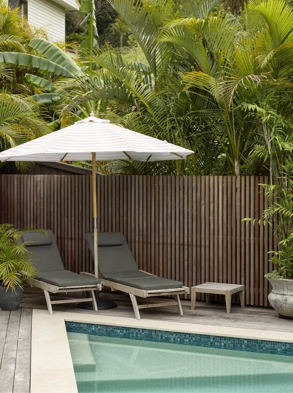 Two lounge chairs with black cushions, a white umbrella, and potted plants near a swimming pool, surrounded by lush green tropical plants and a wooden privacy fence.