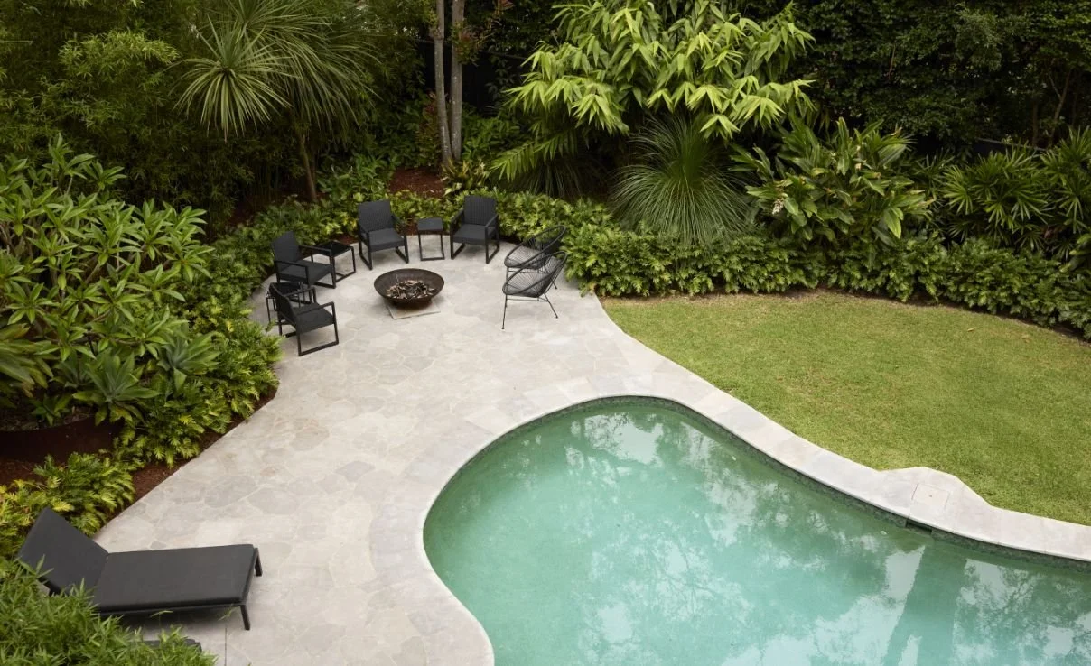 A backyard with a kidney-shaped swimming pool, surrounded by a stone patio and lush green plants, including shrubs, palm trees, and a small grassy area. There are five black chairs arranged in a semi-circle and one lounge chair near the pool.