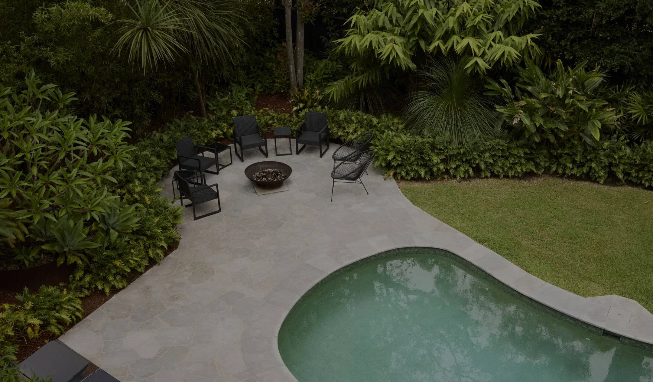 A backyard pool area views from above, featuring a kidney-shaped pool, surrounded by stone-paved patio, green grass, lush bushes, trees, and black outdoor chairs arranged in a semi-circle around a fire pit.