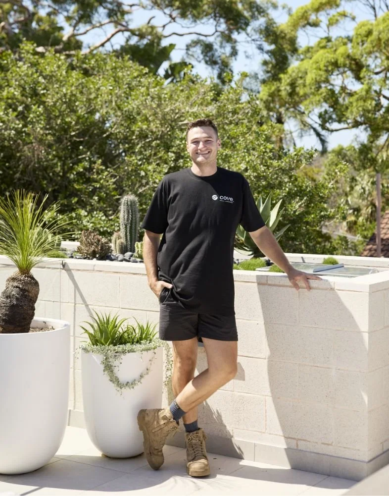 A young man standing outdoors on a sunny day, smiling, wearing a black t-shirt, black shorts, and tan work boots, leaning against a white brick wall surrounded by potted plants and greenery.