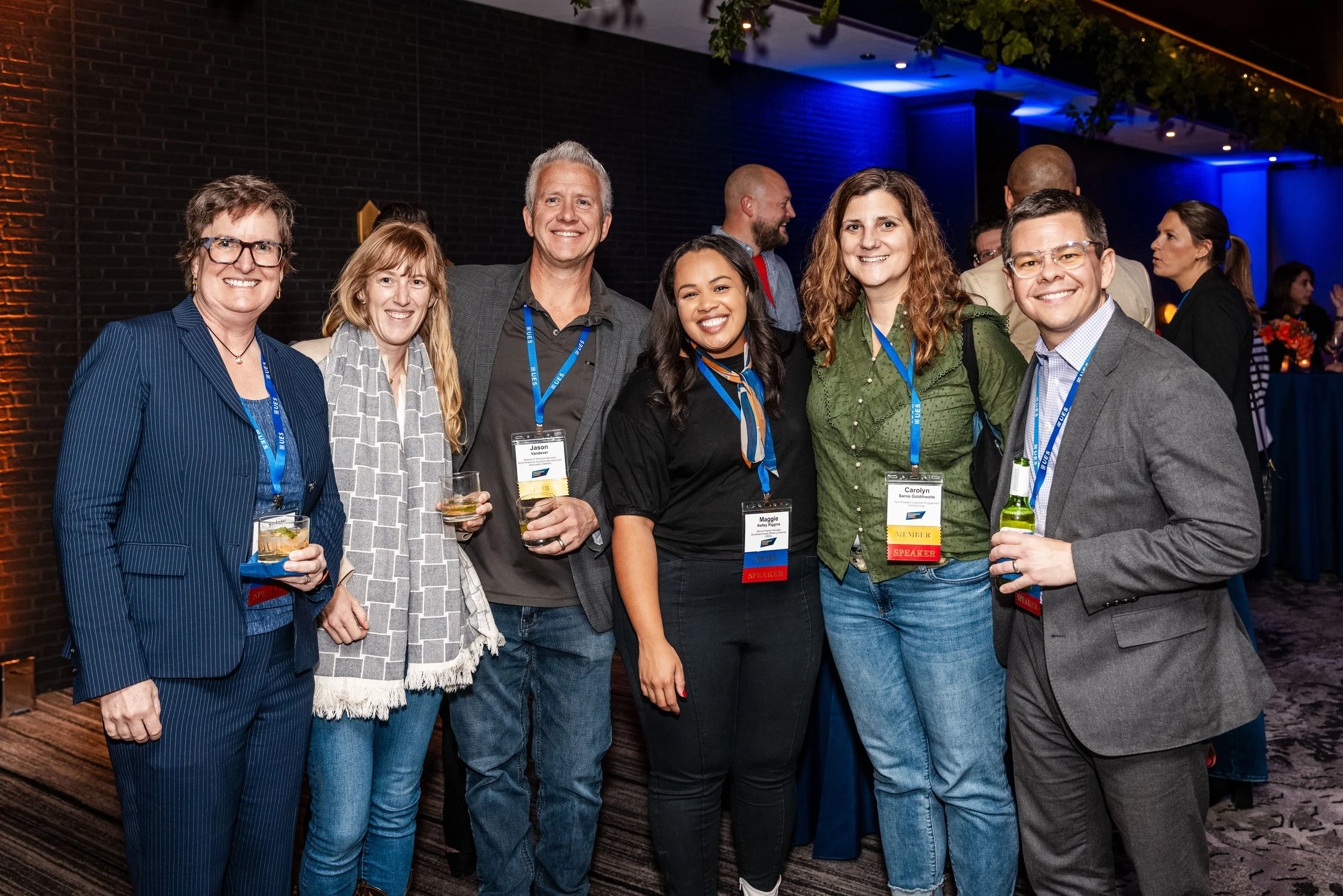 Group of seven diverse people at a professional networking event, smiling and holding drinks, wearing event badges, standing indoors with a dark brick wall and blue lights in the background.