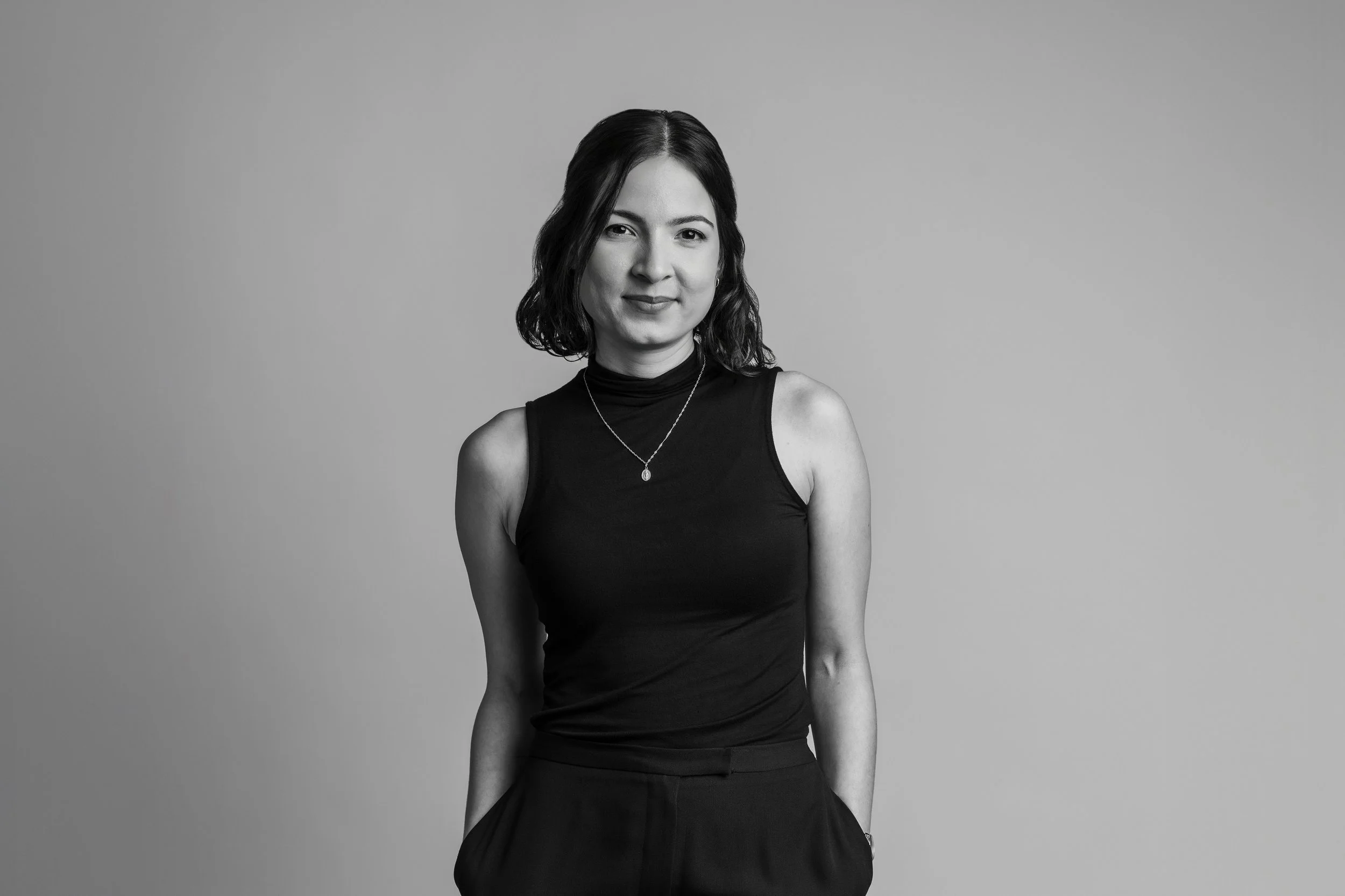 Black and white portrait of a woman with shoulder-length dark hair, wearing a sleeveless black top and a necklace, standing against a plain background.