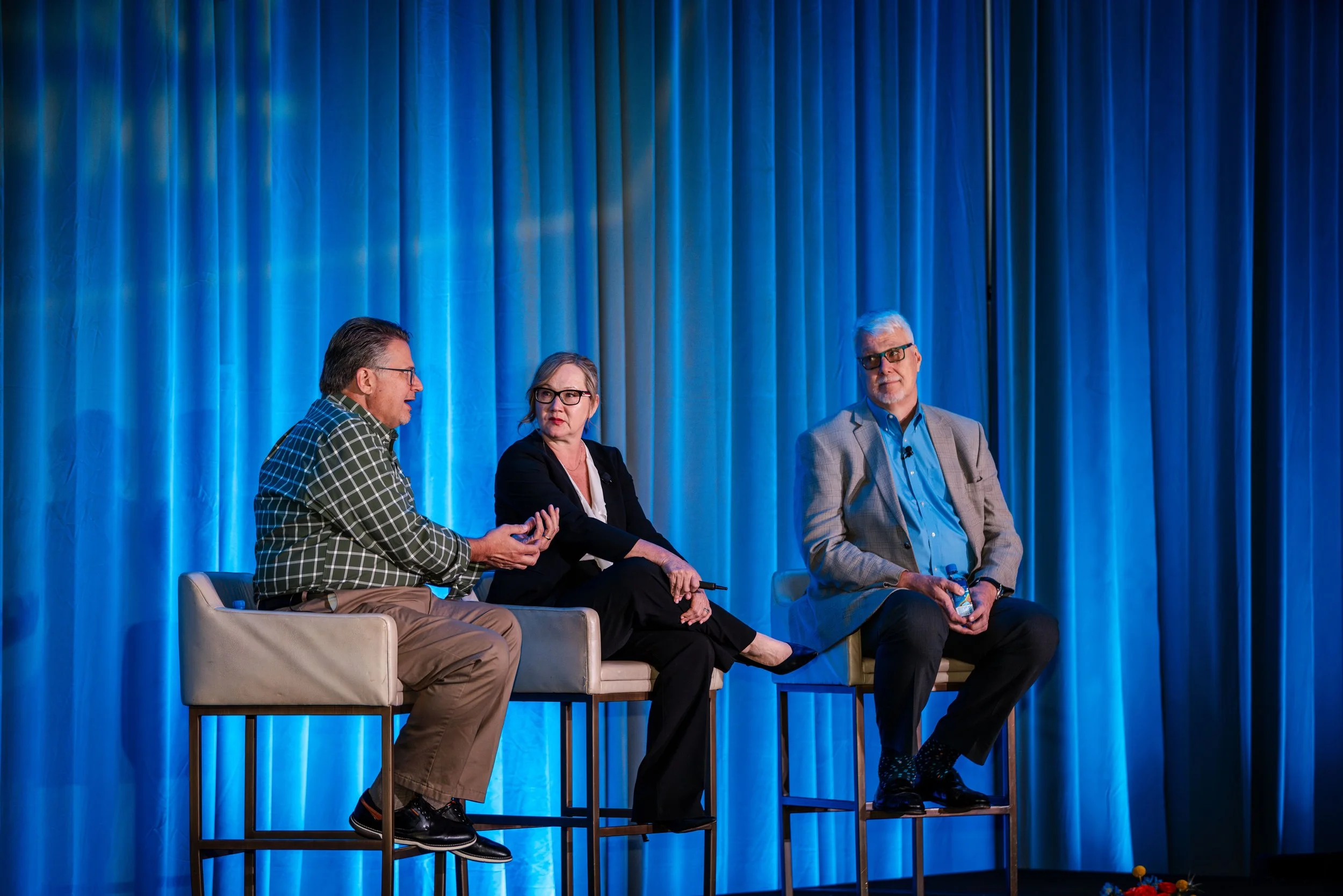 Three people sitting on high chairs on stage, engaged in conversation, with blue curtains as background.