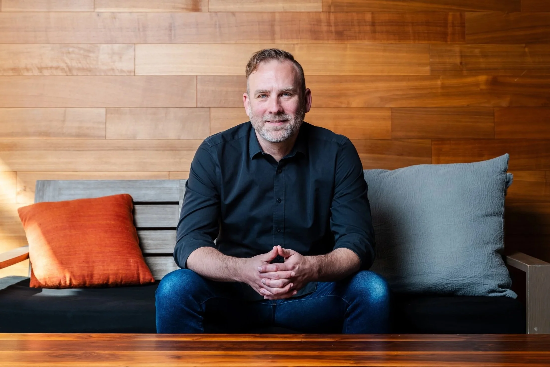A middle-aged man with gray beard and short hair, wearing a black shirt and blue jeans, sitting on a sofa with wooden wall background, orange and gray pillows.