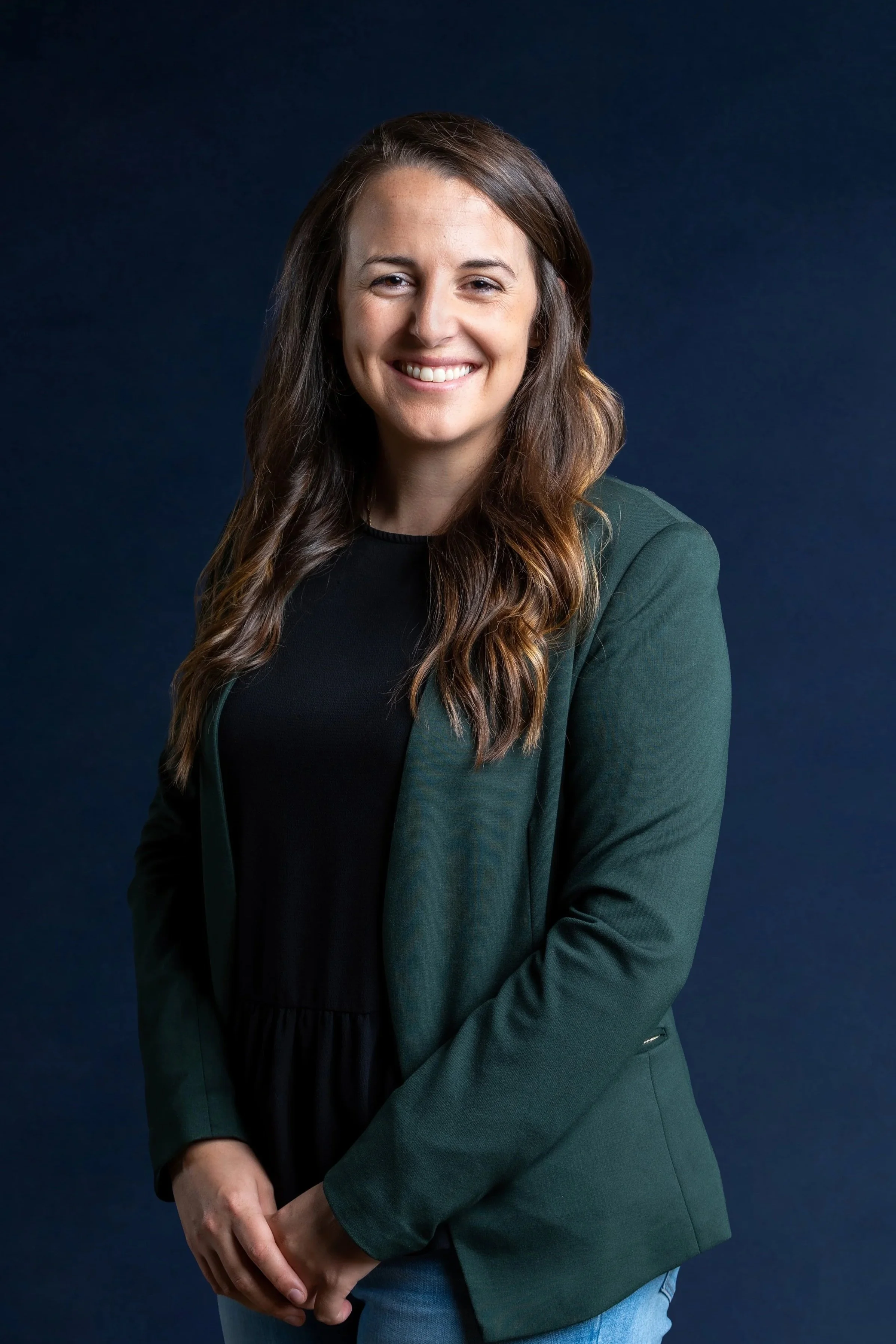 A woman with long brown hair wearing a dark green blazer and black top, smiling against a dark background.