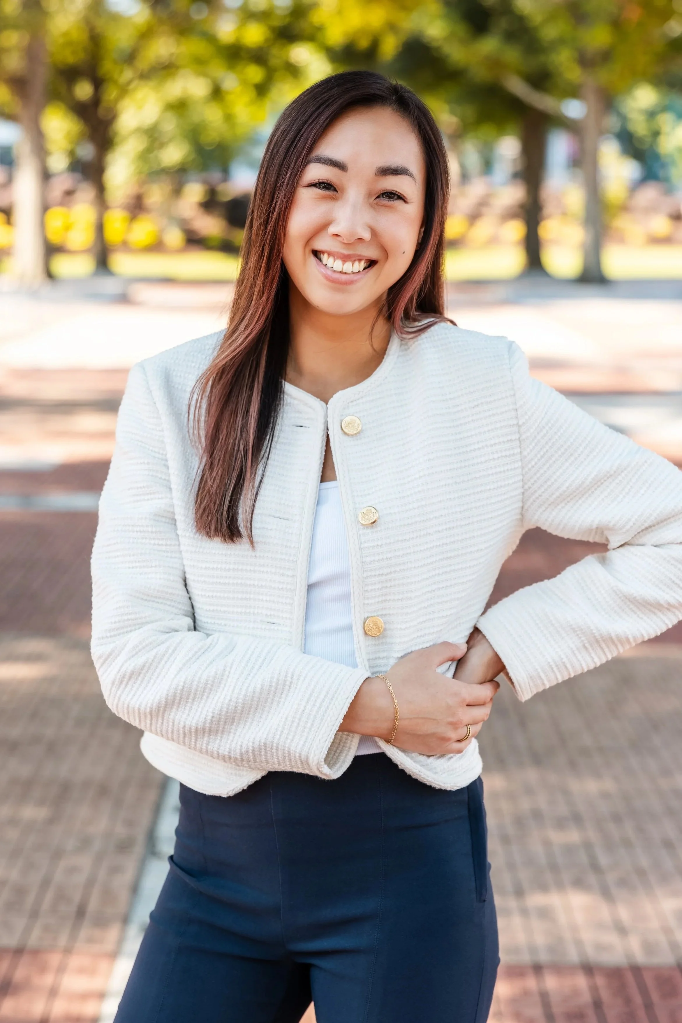 Smiling woman outdoors wearing a white textured jacket and navy pants, with trees and sunlight in the background.
