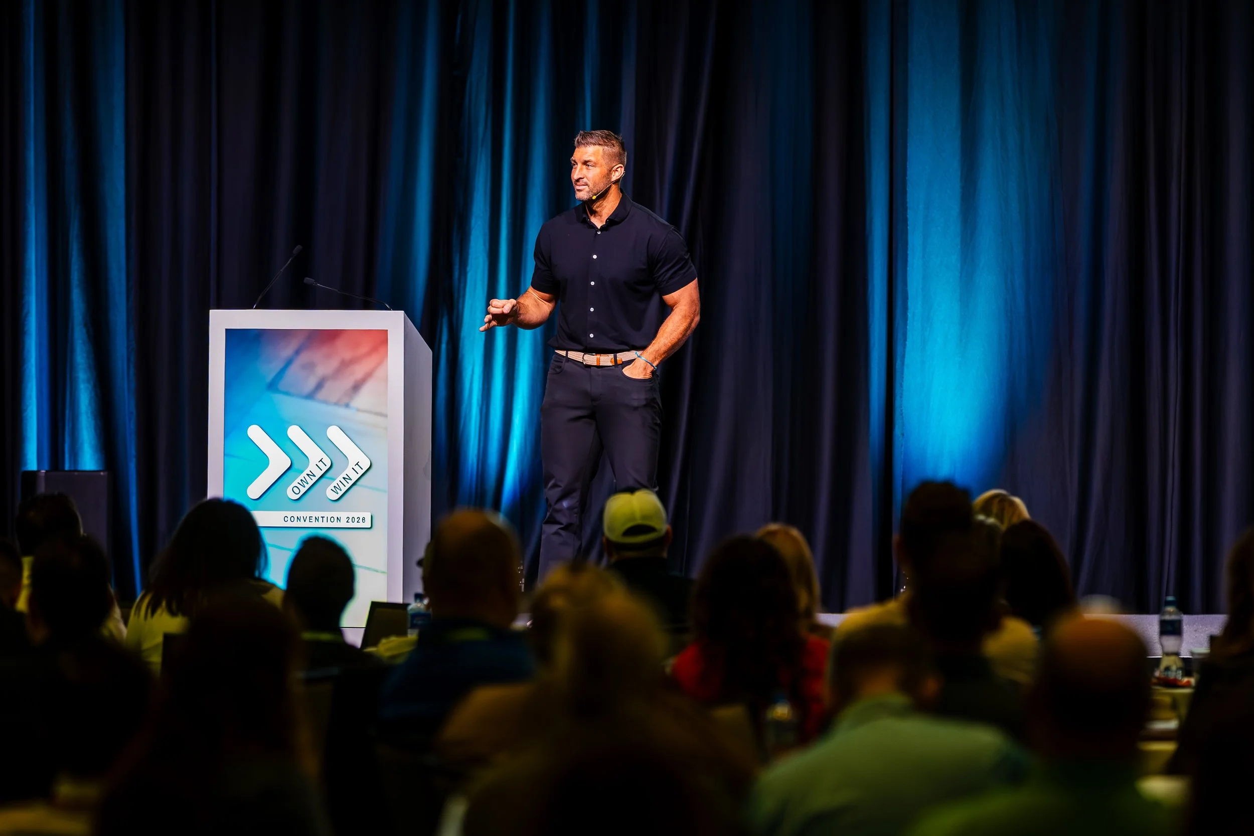 A man giving a presentation at a conference on a stage with a blue curtain backdrop. The lectern displays a logo with arrows and the words 'Own It' and 'Win It,' and a banner below reads 'Convention 2026.' The audience is seated and listening.