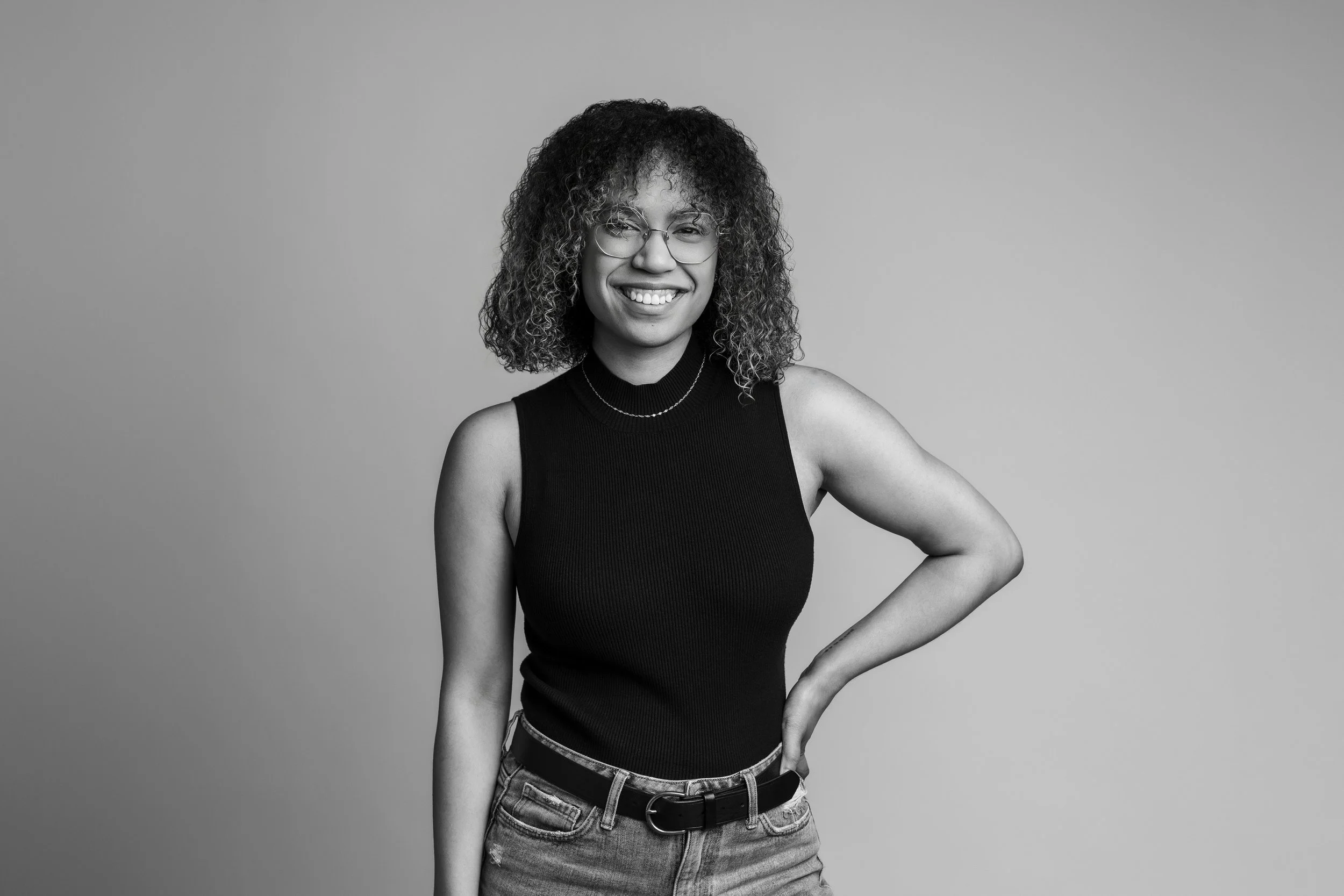 Black and white photo of a woman smiling, wearing glasses, a sleeveless black top, and jeans, standing with one hand on her hip against a plain background.