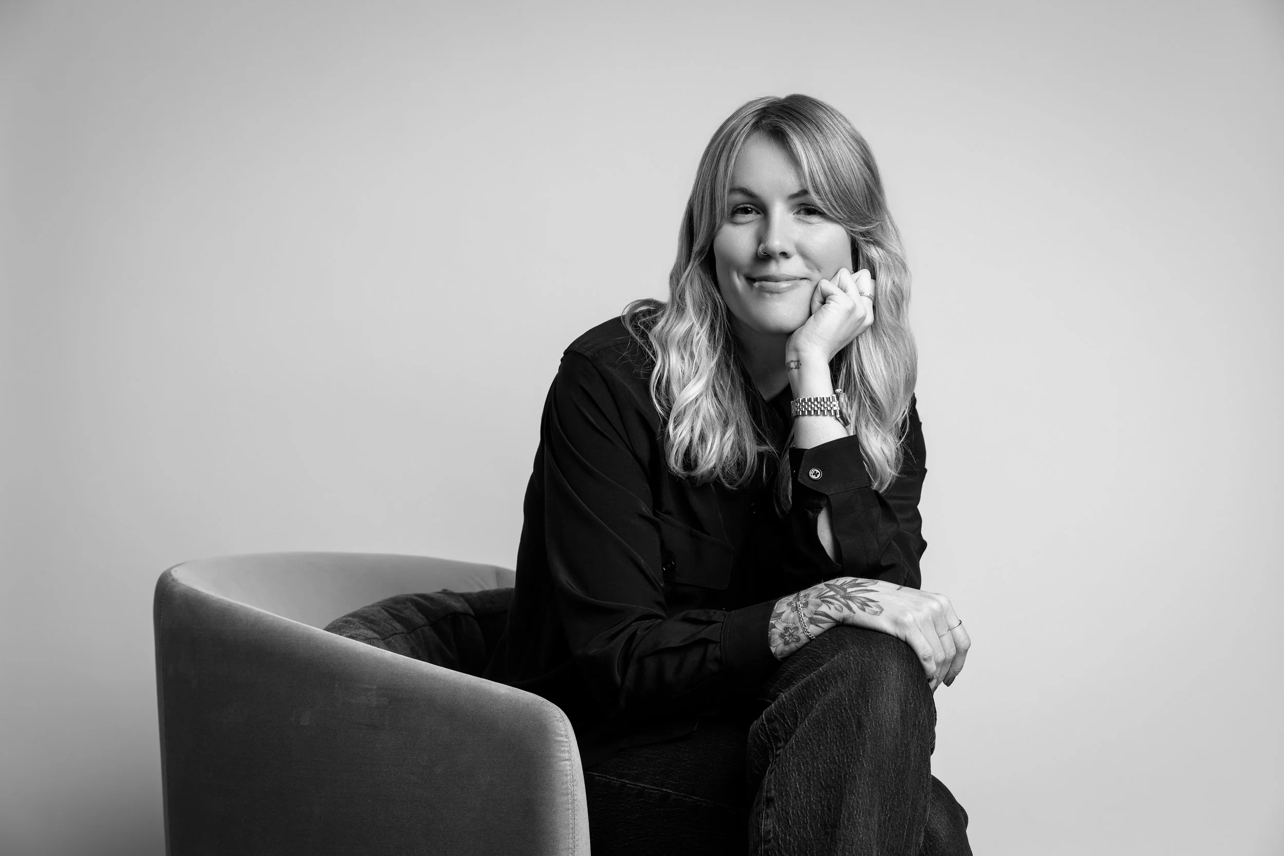 Black and white portrait of a woman sitting on an armchair, smiling, with her chin resting on her hand, wearing a dark shirt and jeans, with visible tattoos on her arm.